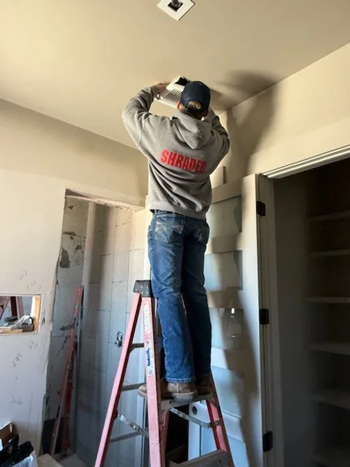 Person standing on a ladder installing a ceiling vent in a room under construction or renovation.