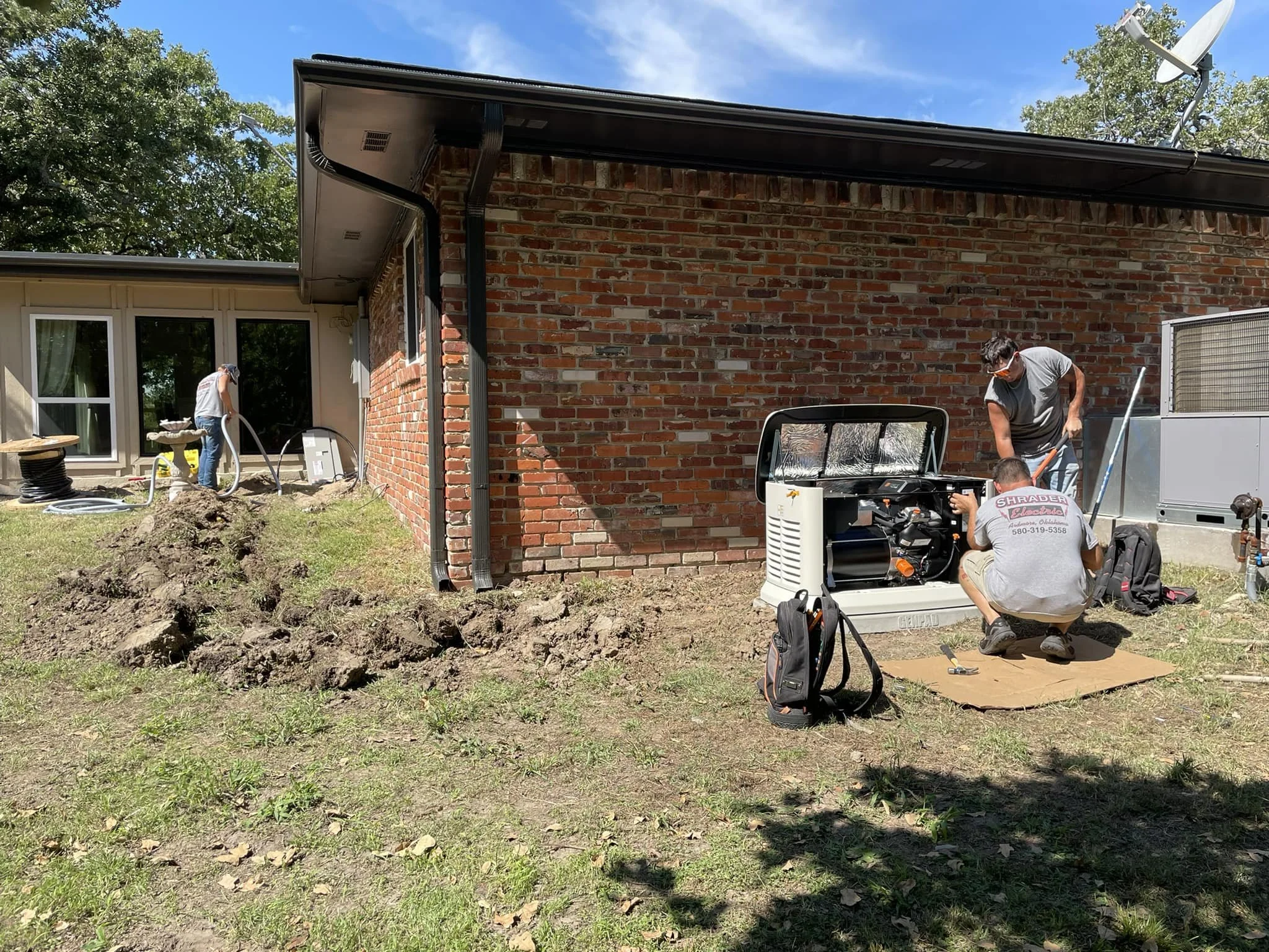 Three workers repairing or servicing an outdoor appliance on a grassy yard beside a brick house. One worker is on the left near some hoses and equipment, while two others are on the right, kneeling and working on a small generator or similar device.