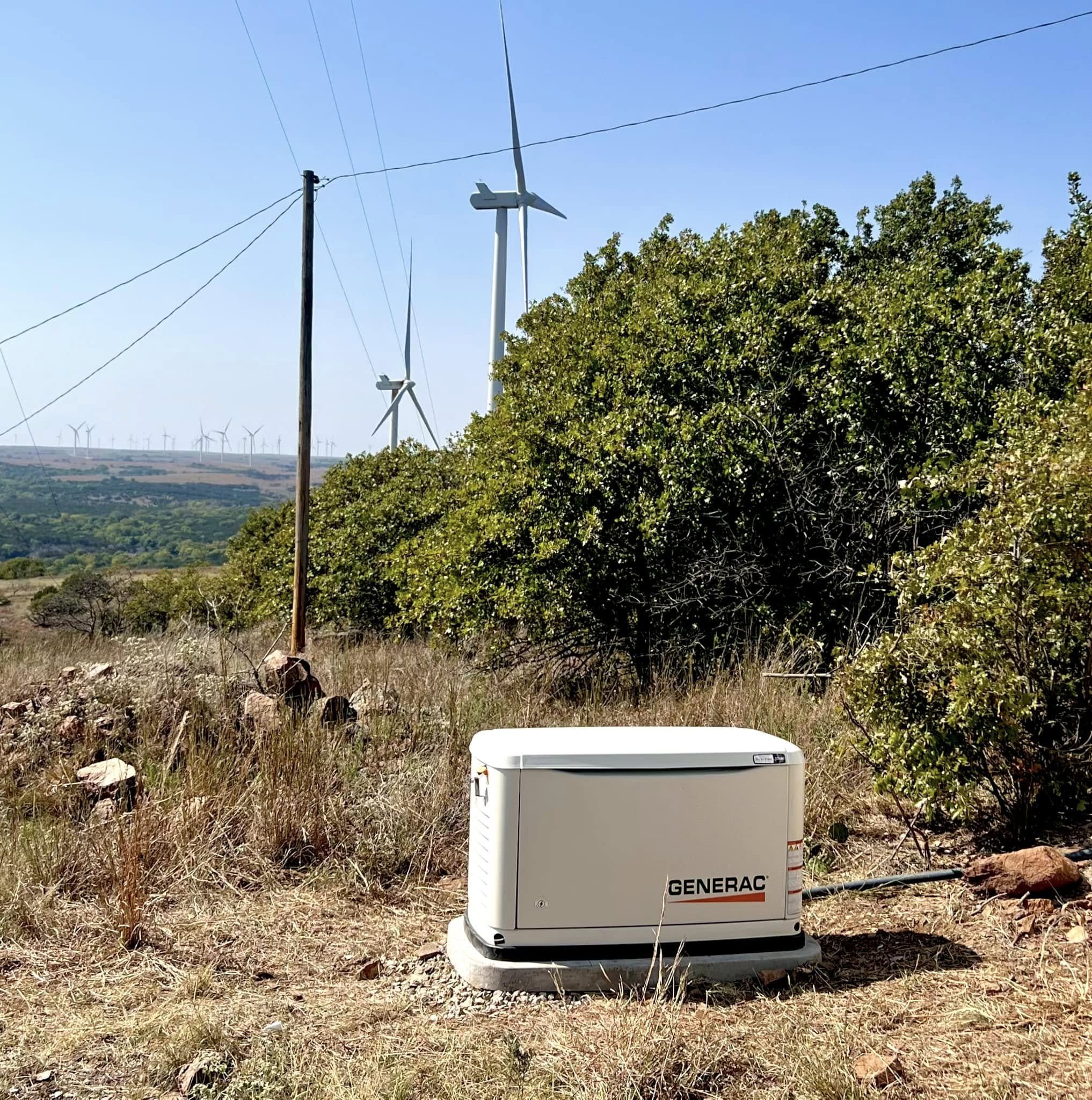 A wind turbine and a Generac generator in a rural landscape with tall grass, bushes, trees, and a clear blue sky.