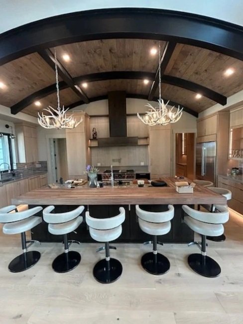 Modern kitchen with a large wooden island, six white bar stools, two chandelier-style light fixtures with antler design, and a vaulted ceiling with wood paneling.