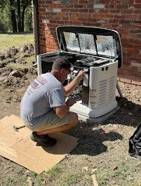 A young man working on repairing or assembling an outdoor gas grill, positioned on a cardboard sheet on grass next to a brick wall.