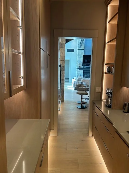 A hallway with wooden cabinets and shelves, leading to a living room with a stone fireplace and a TV, illuminated by natural light.