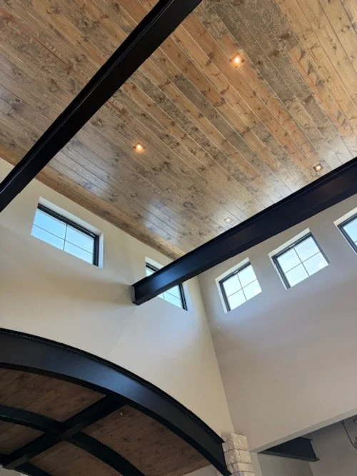 Interior view of a modern building with a wooden ceiling, black beams, and multiple windows letting in natural light.