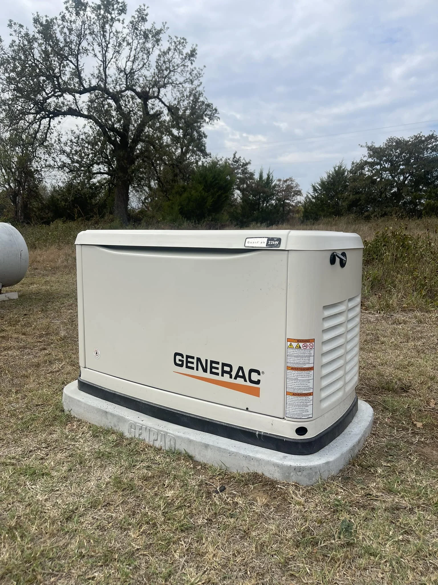 A Generac portable generator installed on a concrete pad in a grassy outdoor area with trees and cloudy sky in the background.