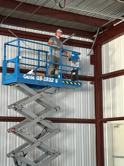 A person standing on a blue Genie GS-1932 scissor lift inside a metal industrial building, wearing safety goggles and a cap.