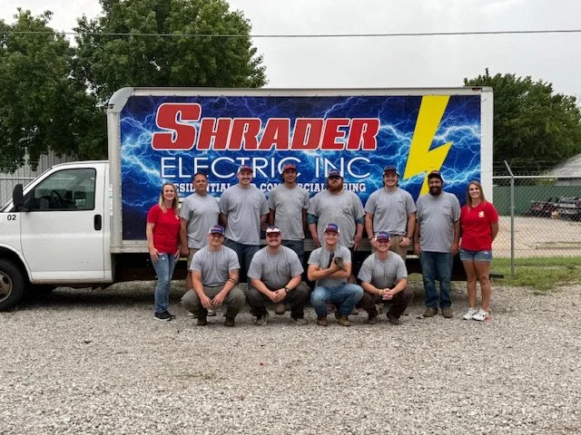 Group of nine people, including five men and four women, standing and kneeling in front of a large truck with a sign that reads 'Shrader Electric Inc.' and a lightning bolt graphic.
