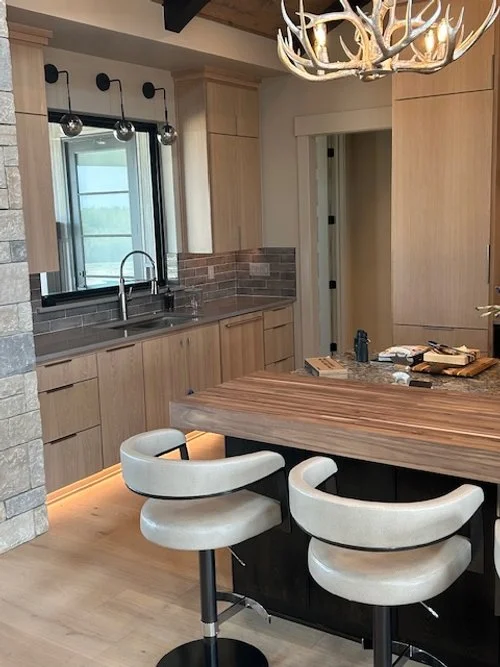 Modern kitchen with light wood cabinets, a gray stone backsplash, a window with black trim, and a kitchen island with two white barstools. An antler chandelier hangs from the ceiling.