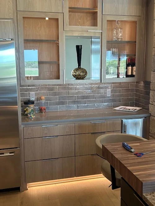Modern kitchen with wooden cabinets, gray brick backsplash, and stainless steel refrigerator. Decor includes a tall black vase with a gold patterned base, wine bottles, and glasses. There is a countertop with jars and a magazine, and a dining table w