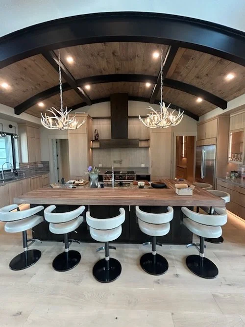 Modern kitchen with a large wooden island, six white swivel bar stools, antler chandeliers, and a vaulted ceiling with wood paneling.