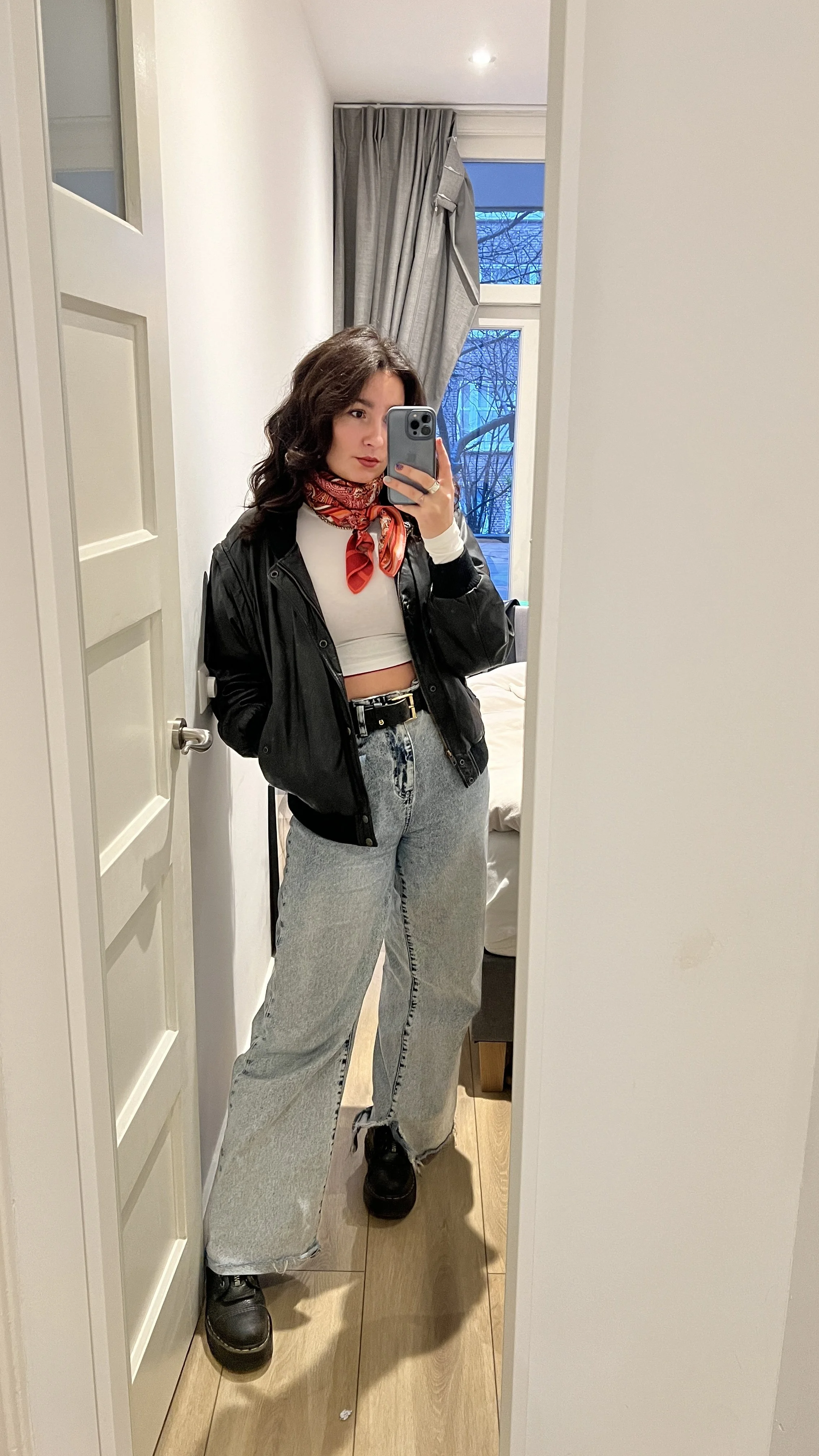 Eine junge Frau macht ein Selfie in einem Spiegel, trägt eine schwarze Jacke, ein weißes Crop-Top, große Jeans, schwarze Stiefel und einen roten Schal.
