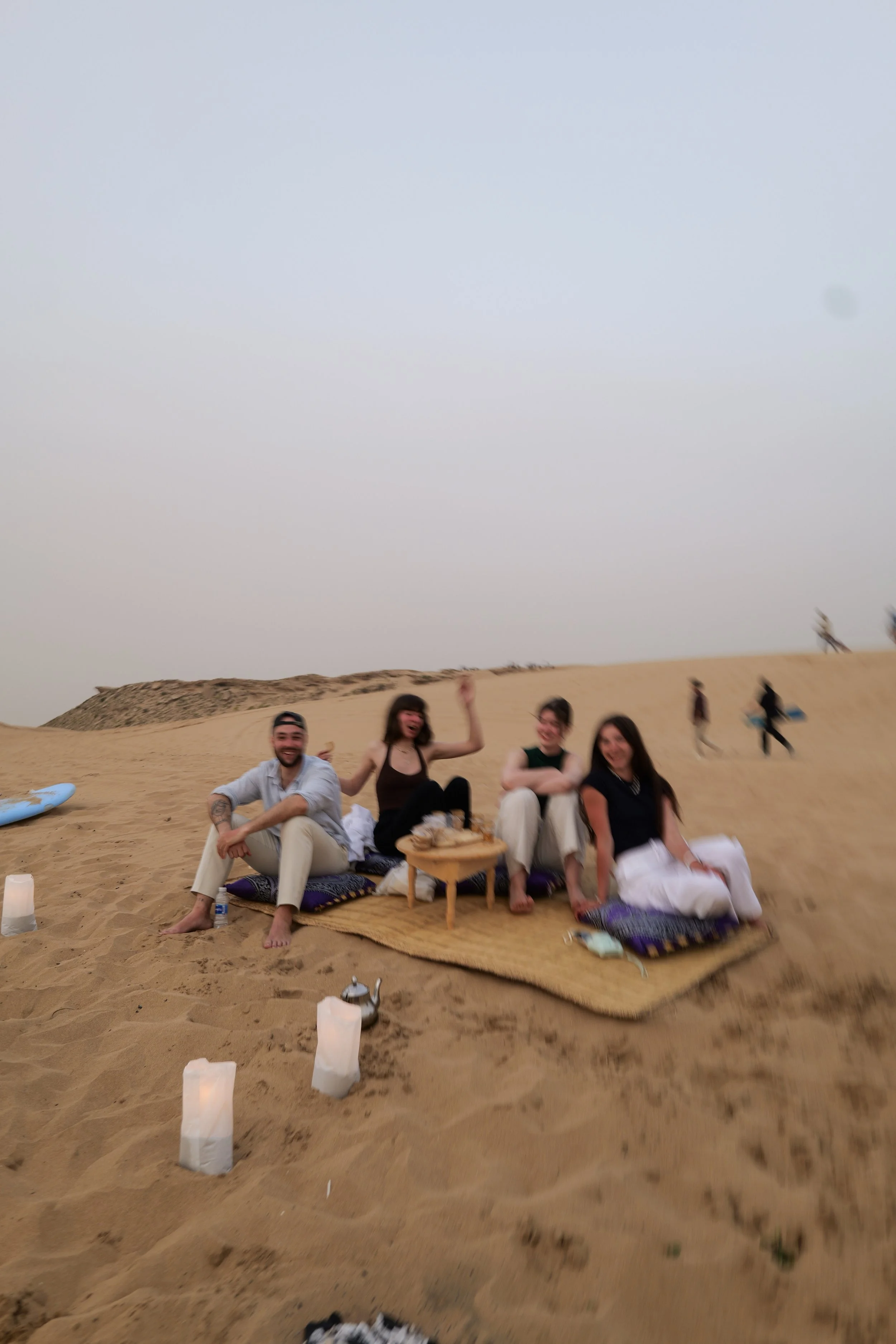 Gruppen von Menschen, die auf einer Sanddüne im Sonnenuntergang sitzen und lachen, mit Tee und Kerzen im Licht.