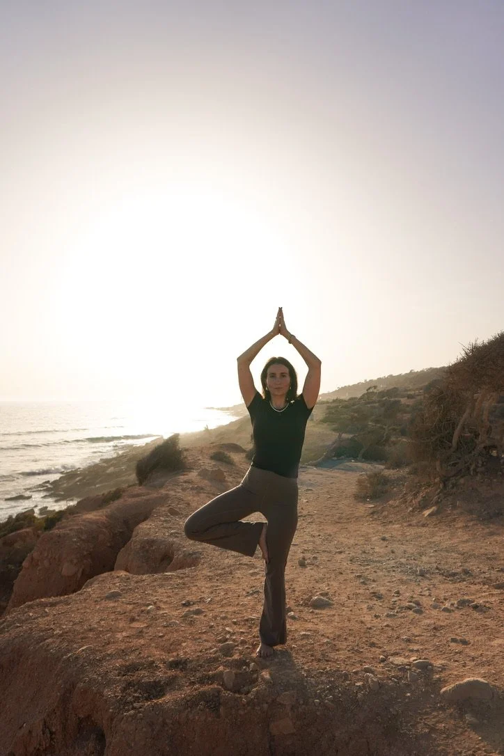 Frau beim Yoga am Strand, Sonnenuntergang, Felsen und Meer im Hintergrund.