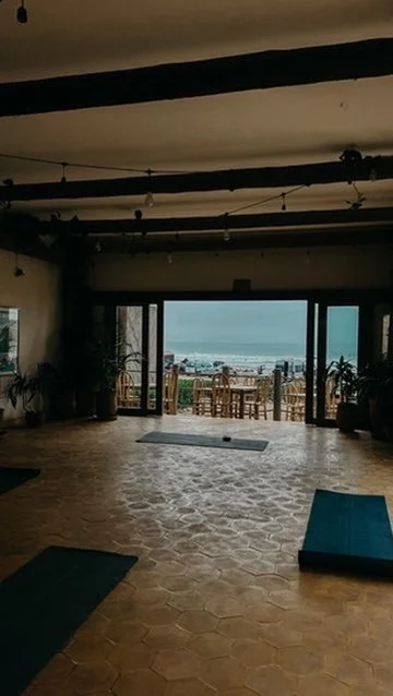 Innenraum eines Raums, Blick auf eine Terrasse mit Stühlen und Tisch, Blick aufs Meer durch große Glasfenster.