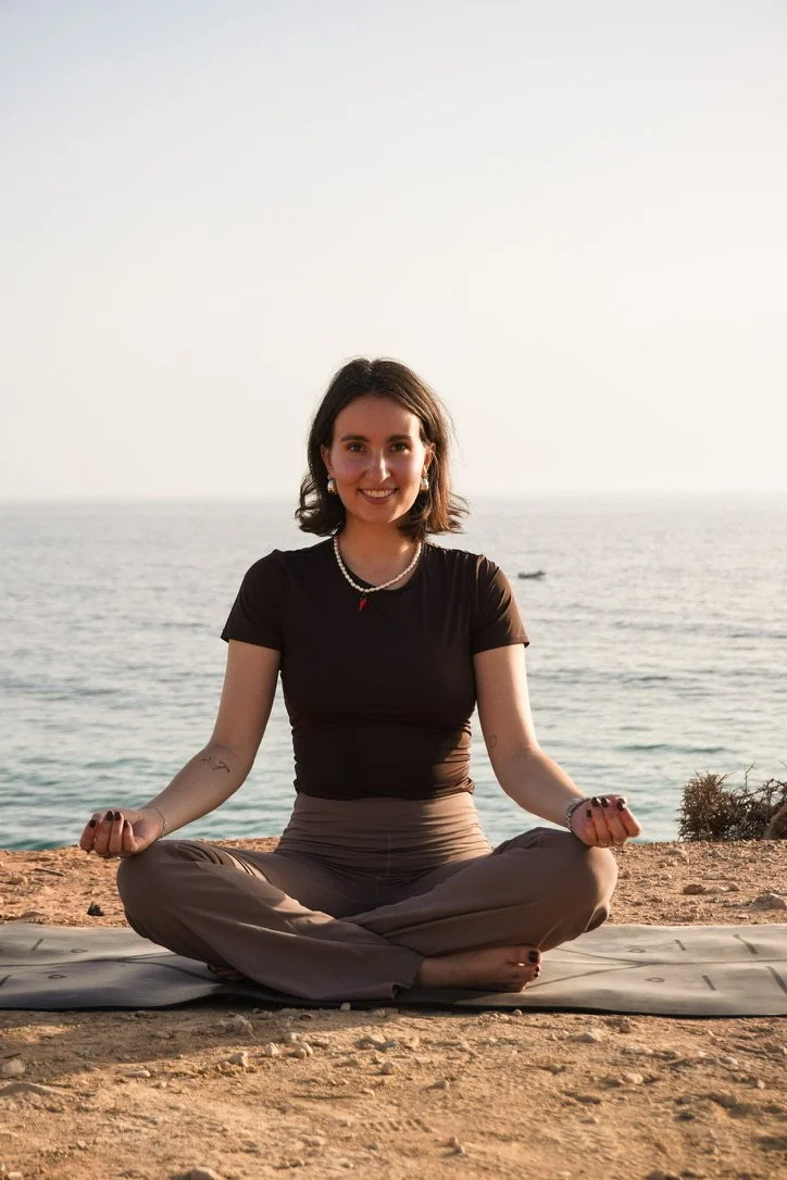 Frau, die auf einem Yoga-Matten am Strand im Schneidersitz meditiert, mit Blick auf das Meer im Hintergrund bei Sonnenuntergang.