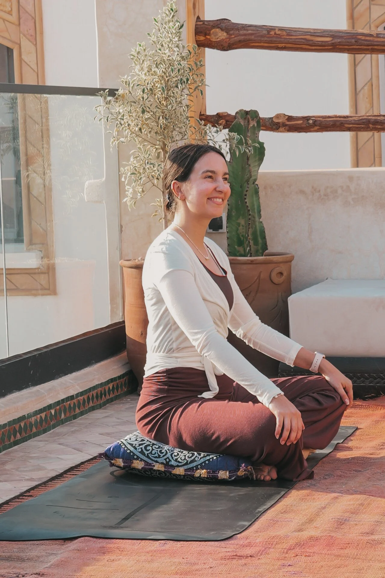 Eine Frau sitzt auf einem Meditationskissen im Innenraum, umgeben von Pflanzen und Holzdekorationen, während sie in einer Meditationshaltung entspannt aussieht.