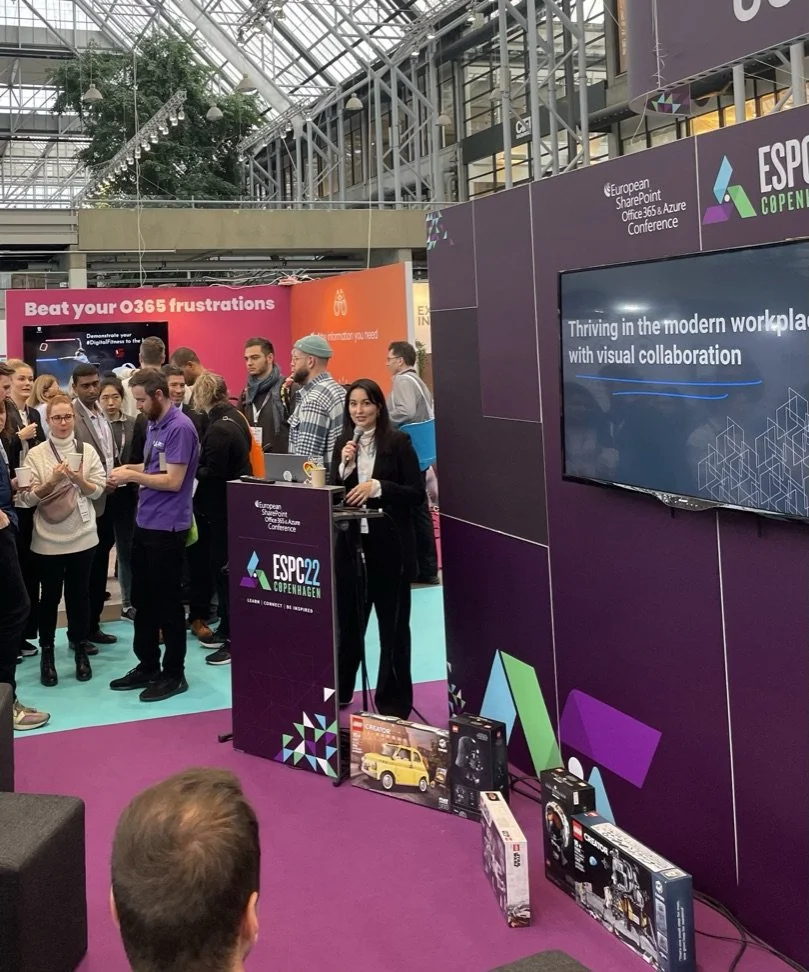 Eine Messehalle mit mehreren Menschen, die an einem Stand der ECSP22 Conference in Kopenhagen teilnehmen. Es gibt eine Frau, die an einem Podium steht und eine Präsentation hält, während andere Personen zuhören. Es sind verschiedene Produkte auf dem Boden um den Stand herum, darunter Spielzeugautos und Elektronikartikel.