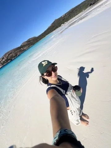 Person am Strand, Sonnenbrille, Hut, Blick auf die Kamera, klarer Himmel, sandiger Strand, Wasser, Hügel im Hintergrund.