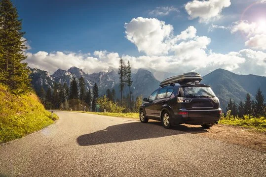 SUV with a roof cargo box parked on a mountain road with trees and mountain peaks in the background.