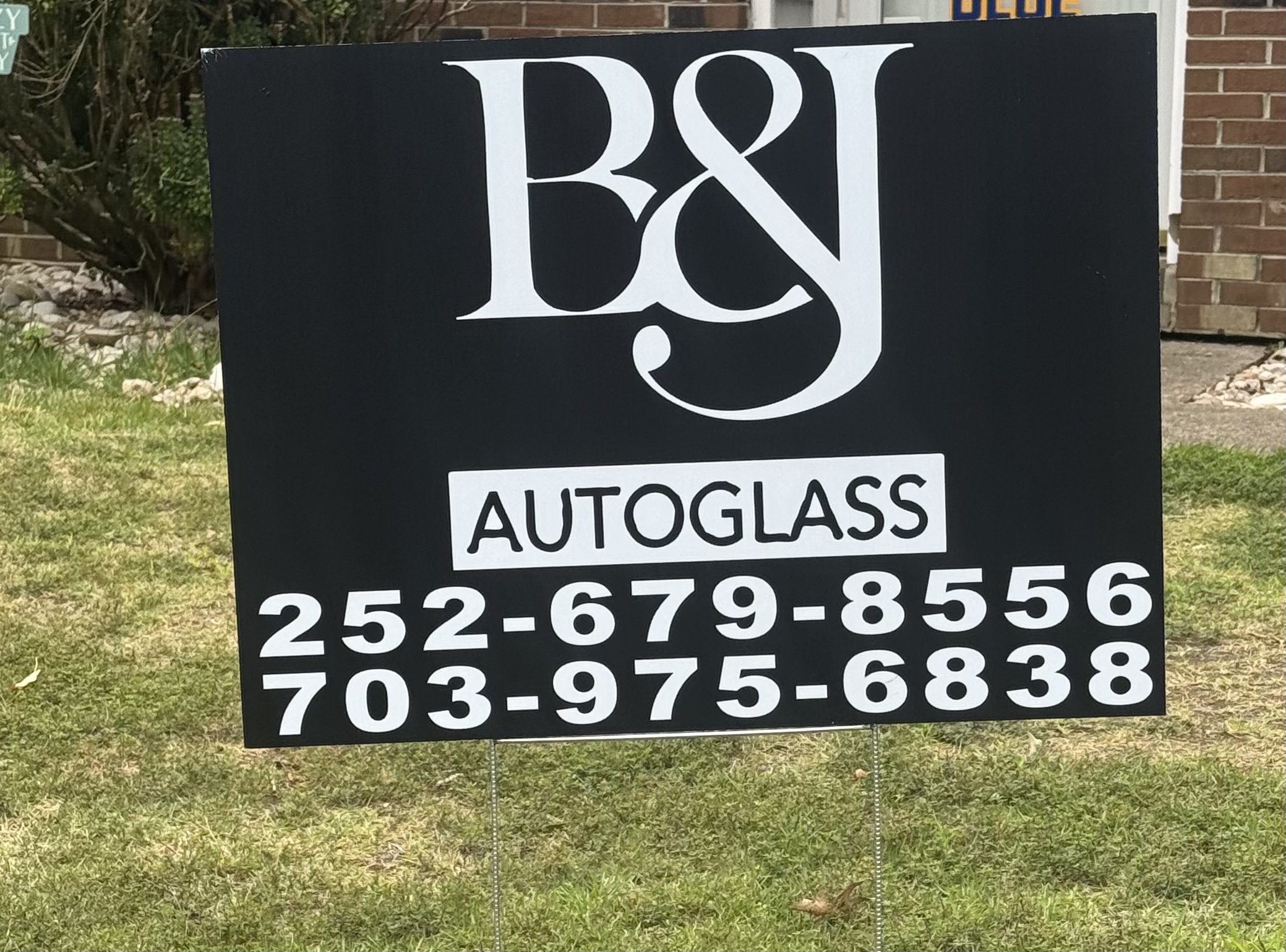 A black and white sign for a business named R&J Autoglass, displaying two phone numbers: 252-679-8556 and 703-975-6838, in a grassy outdoor area with some bushes and a brick building in the background.