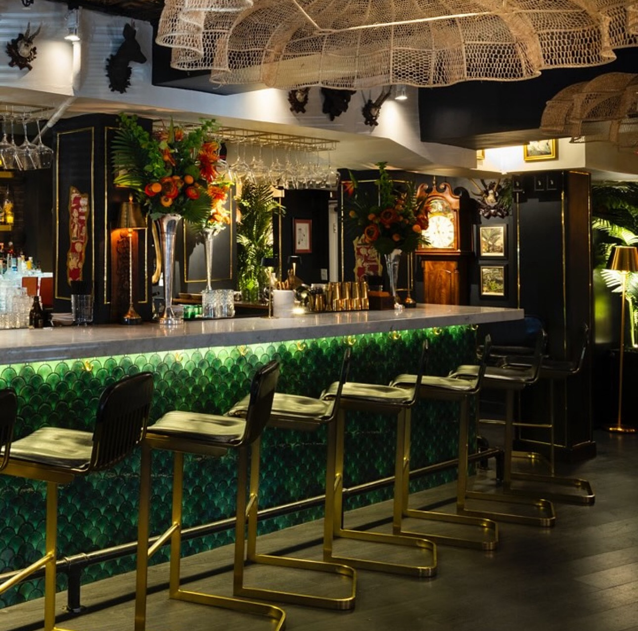 A stylish bar area with black and gold accents, green tiled bar front, floral arrangements, and hanging glassware.