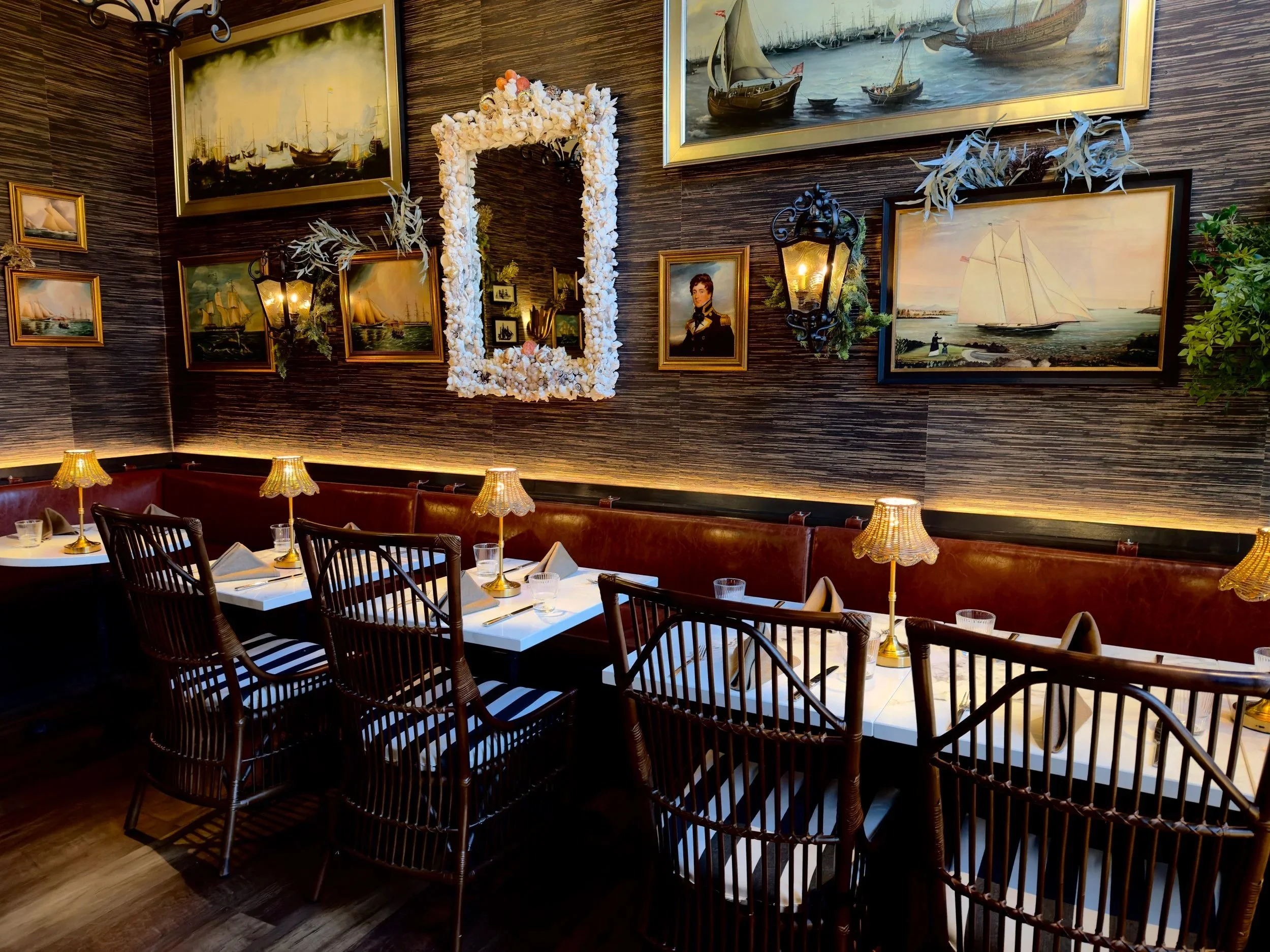 A cozy restaurant with a nautical theme decor featuring a wall decorated with various framed paintings of ships and seascapes, a large mirror with a shell frame, and wall-mounted lamps. There is a long white table with wooden chairs, set with table lamps, glasses, and napkins.