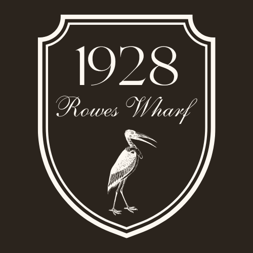 A black shield-shaped logo with white borders displaying the year '1928', the text 'Rowes Wharf', and an illustration of a heron standing with its beak open.