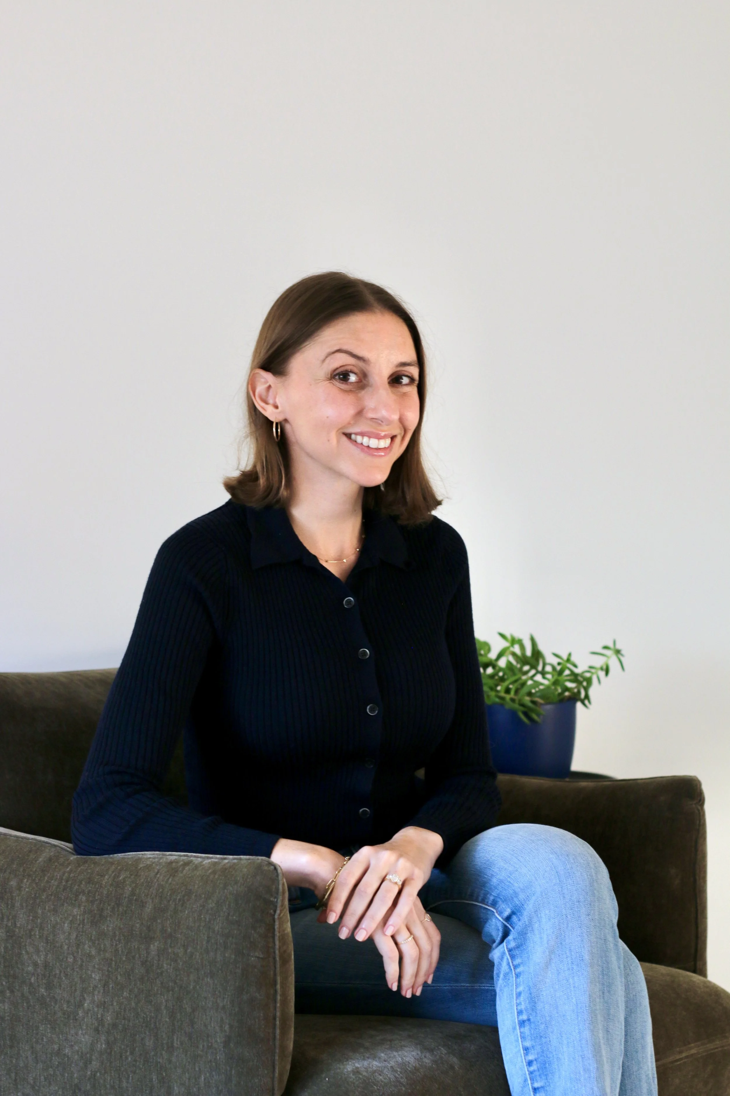 A woman with shoulder-length brown hair, wearing a navy blue sweater and light blue jeans, sitting on a gray couch with her hands resting on her lap, smiling in a room with a white wall and a potted plant in a blue pot.