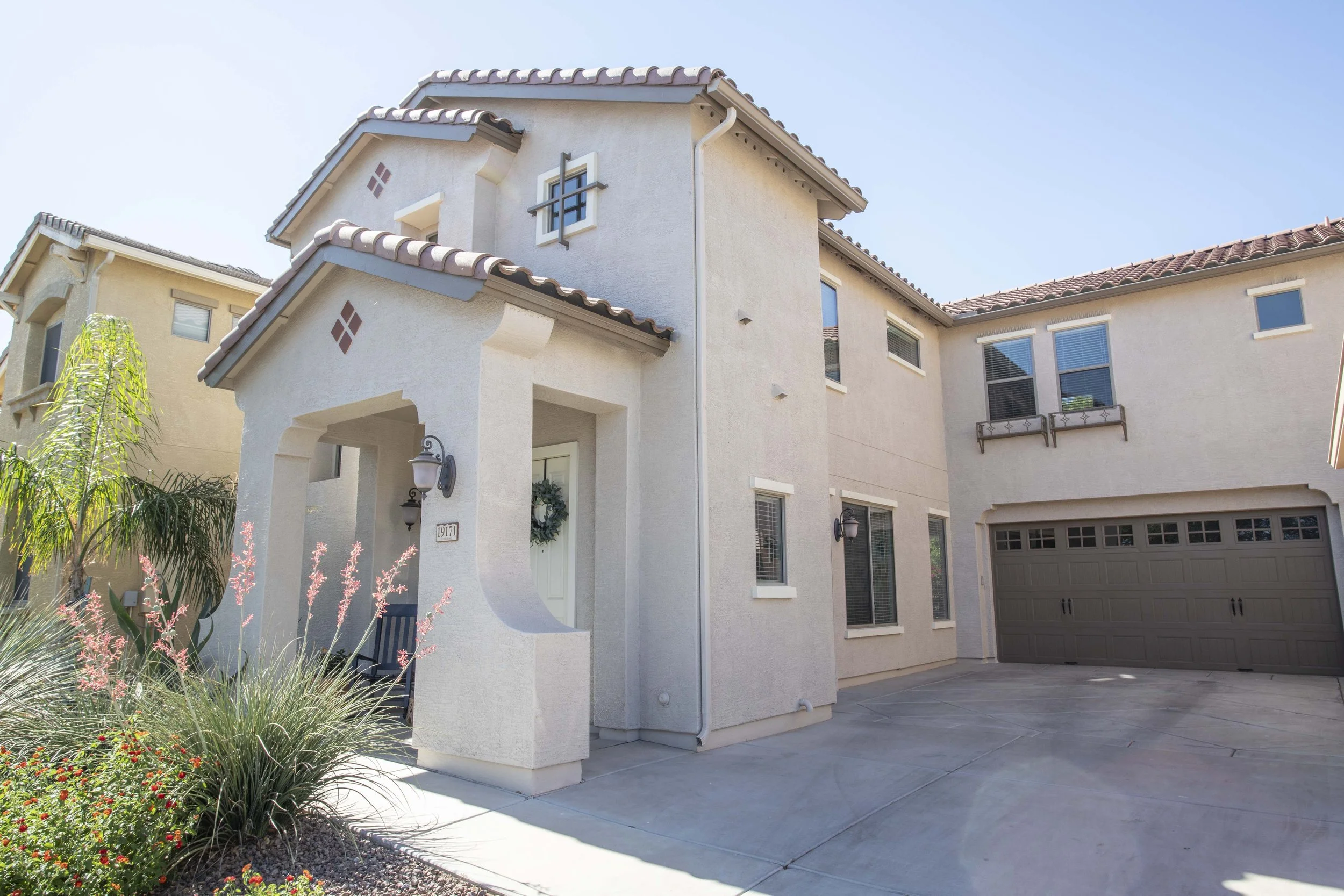 Front view of a two-story beige house with a tiled roof, featuring a garage, front porch with plants and a wreath on the door, and multiple windows under a clear blue sky.