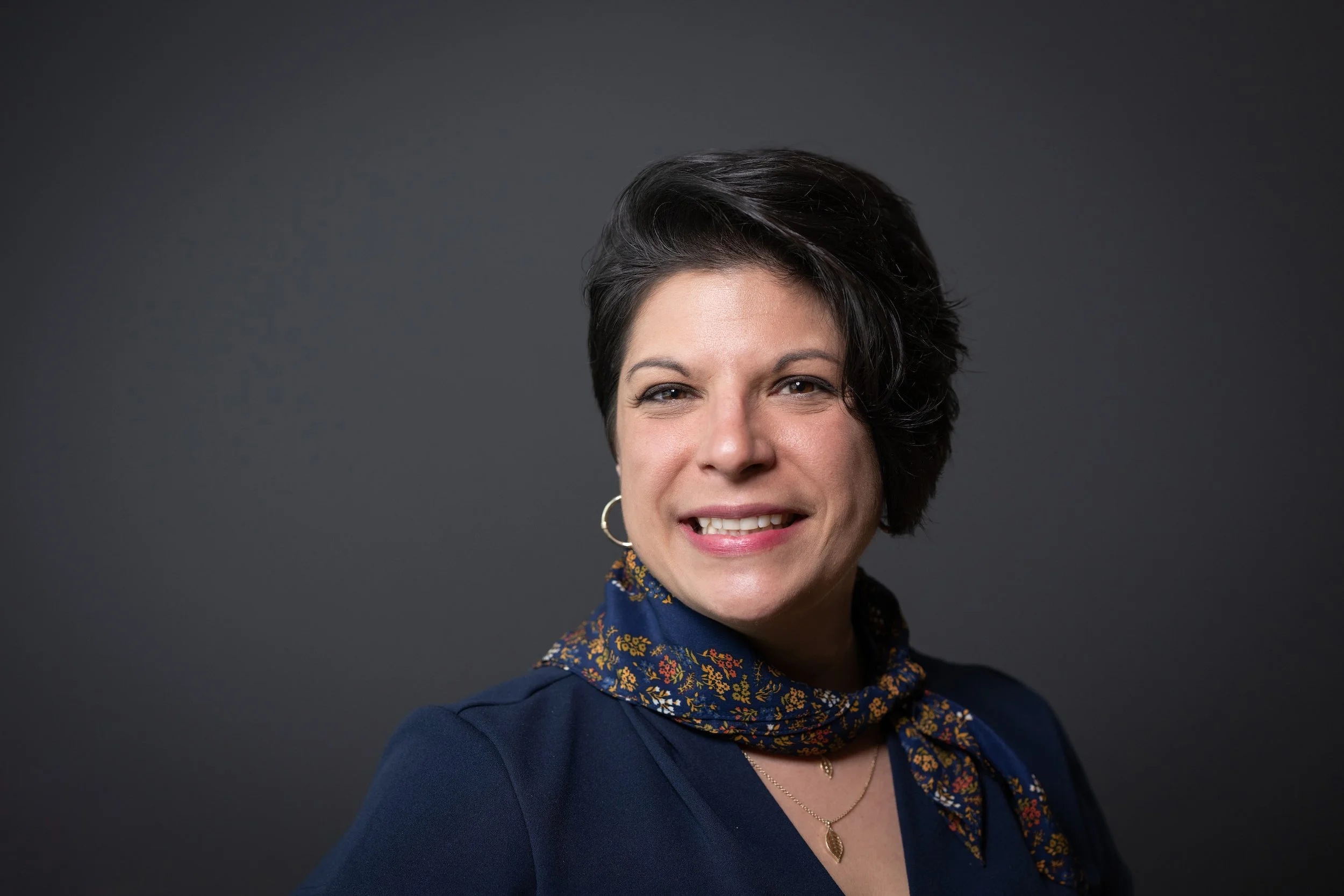 A woman with short dark hair, wearing a navy blue top, a floral scarf, hoop earrings, and layered necklaces, smiling at the camera against a dark grey background.