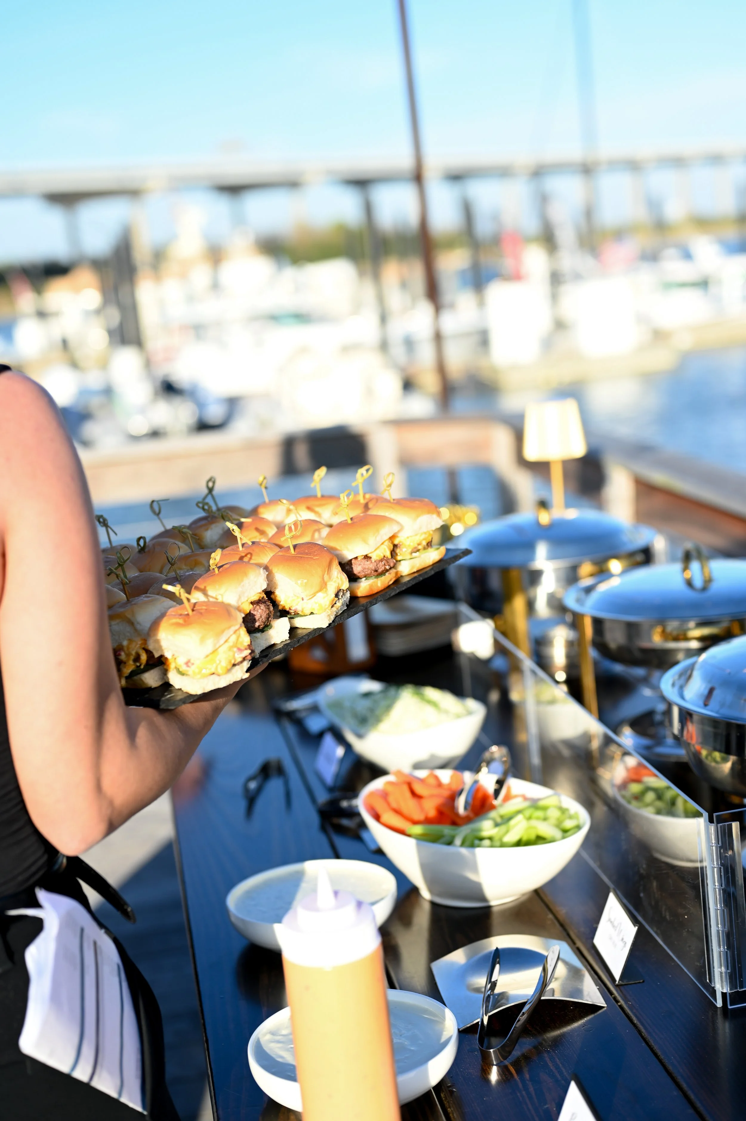 A person holding a tray of small sandwiches with sliders at an outdoor party near a marina with boats and a bridge in the background.