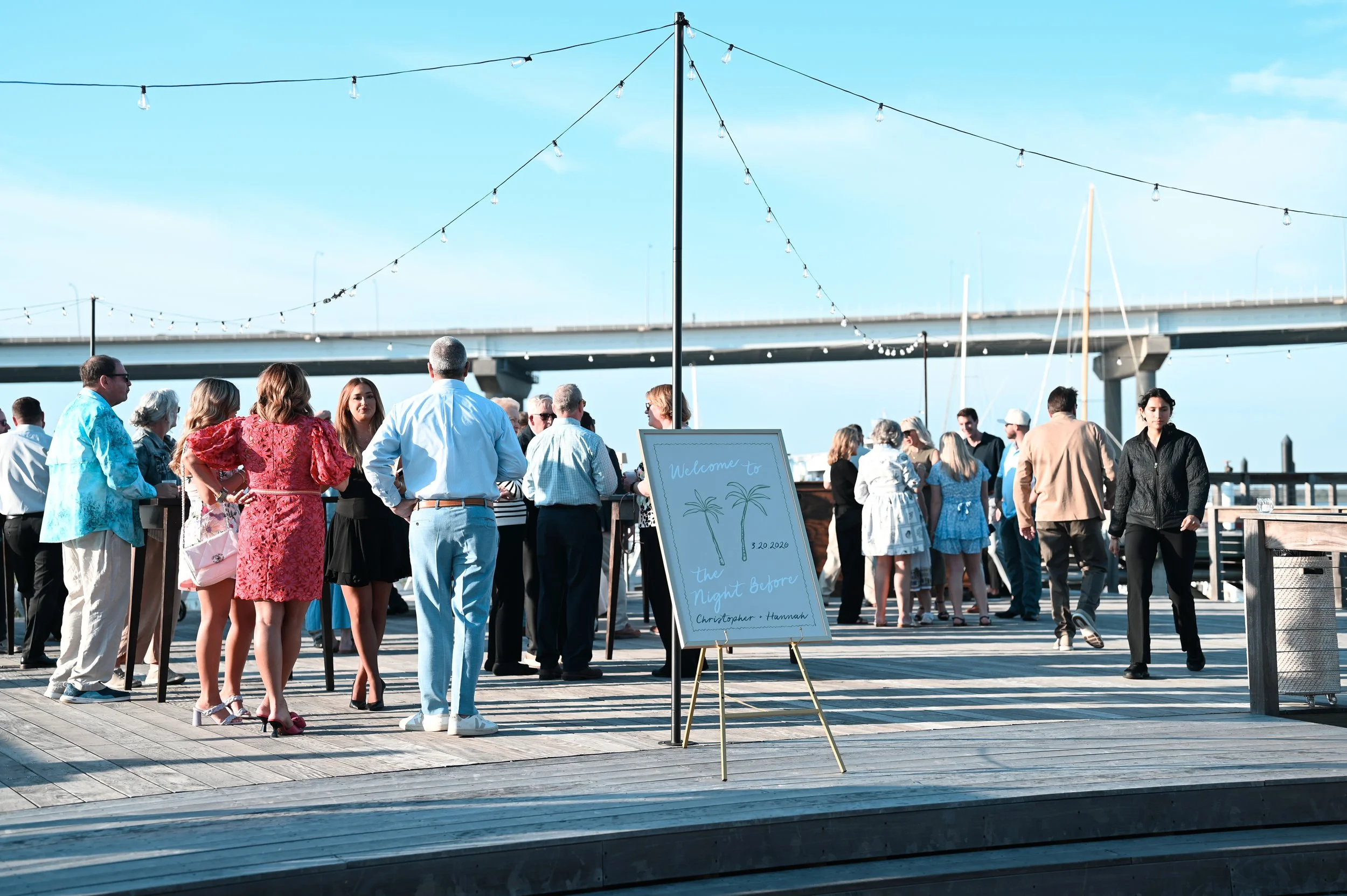 People gathered at an outdoor area with string lights, near a bridge, with a sign that reads "Welcome to the Night Before, Christopher + Hannah, 3.20.2020." The sky is blue and the scene suggests a social gathering or event.