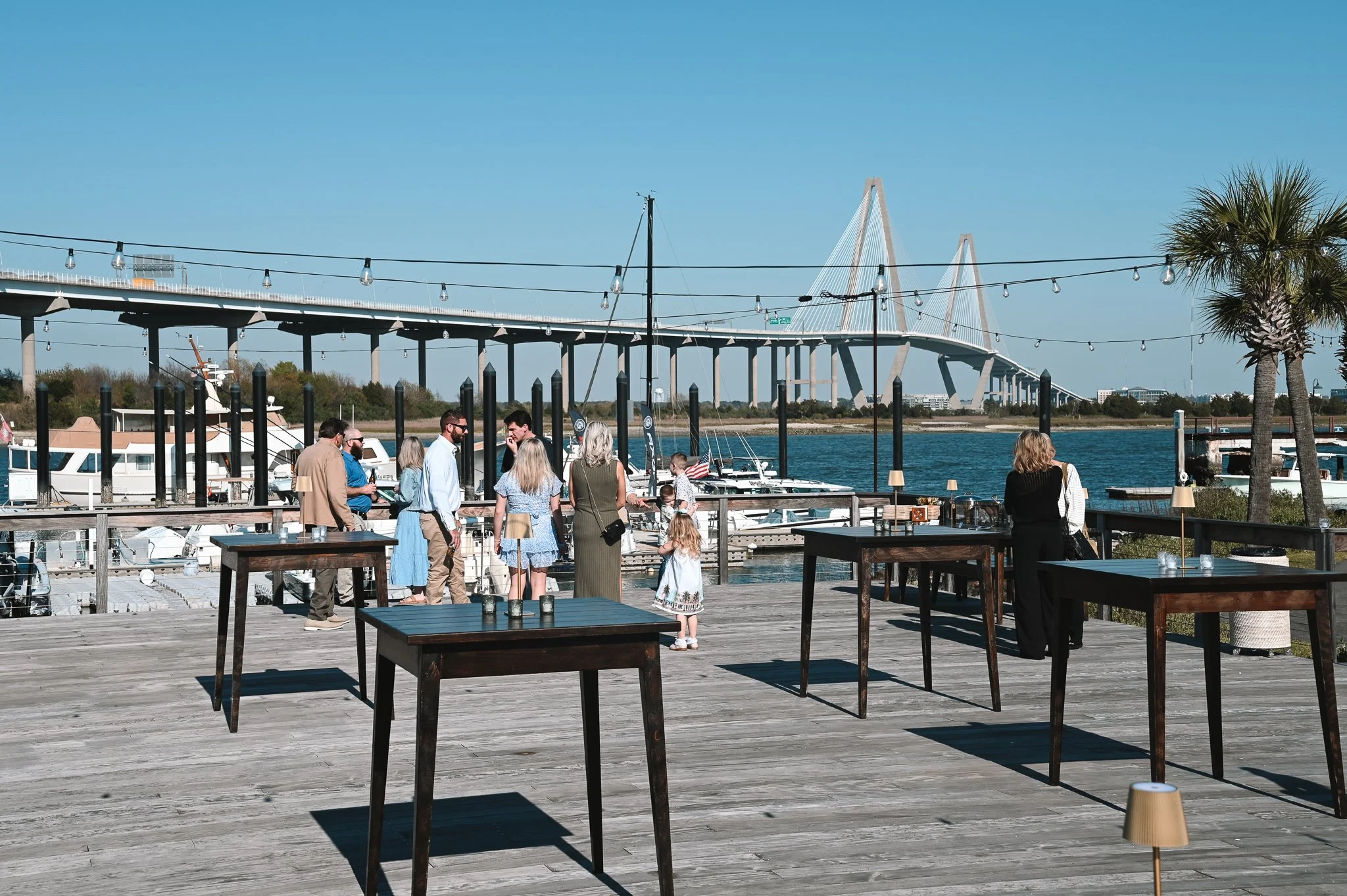 People gathered on a wooden dock enjoying a sunny day near boats and water, with a modern bridge in the background and palm trees on the right.