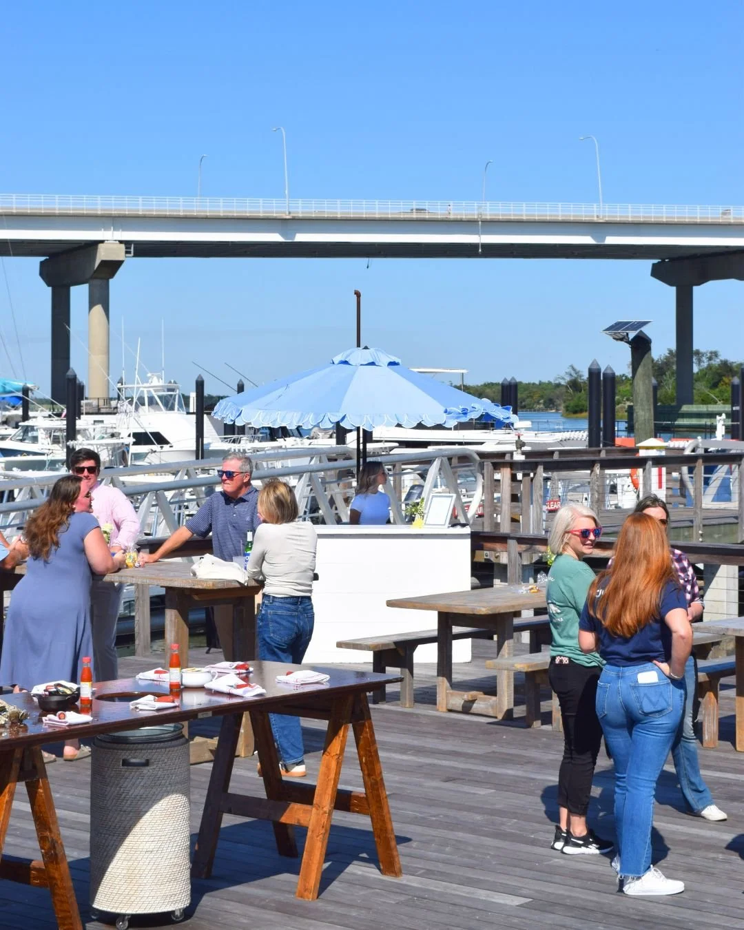 People gathering and chatting on a wooden dock area with outdoor tables and a large blue umbrella, with boats and a bridge in the background on a clear, sunny day.