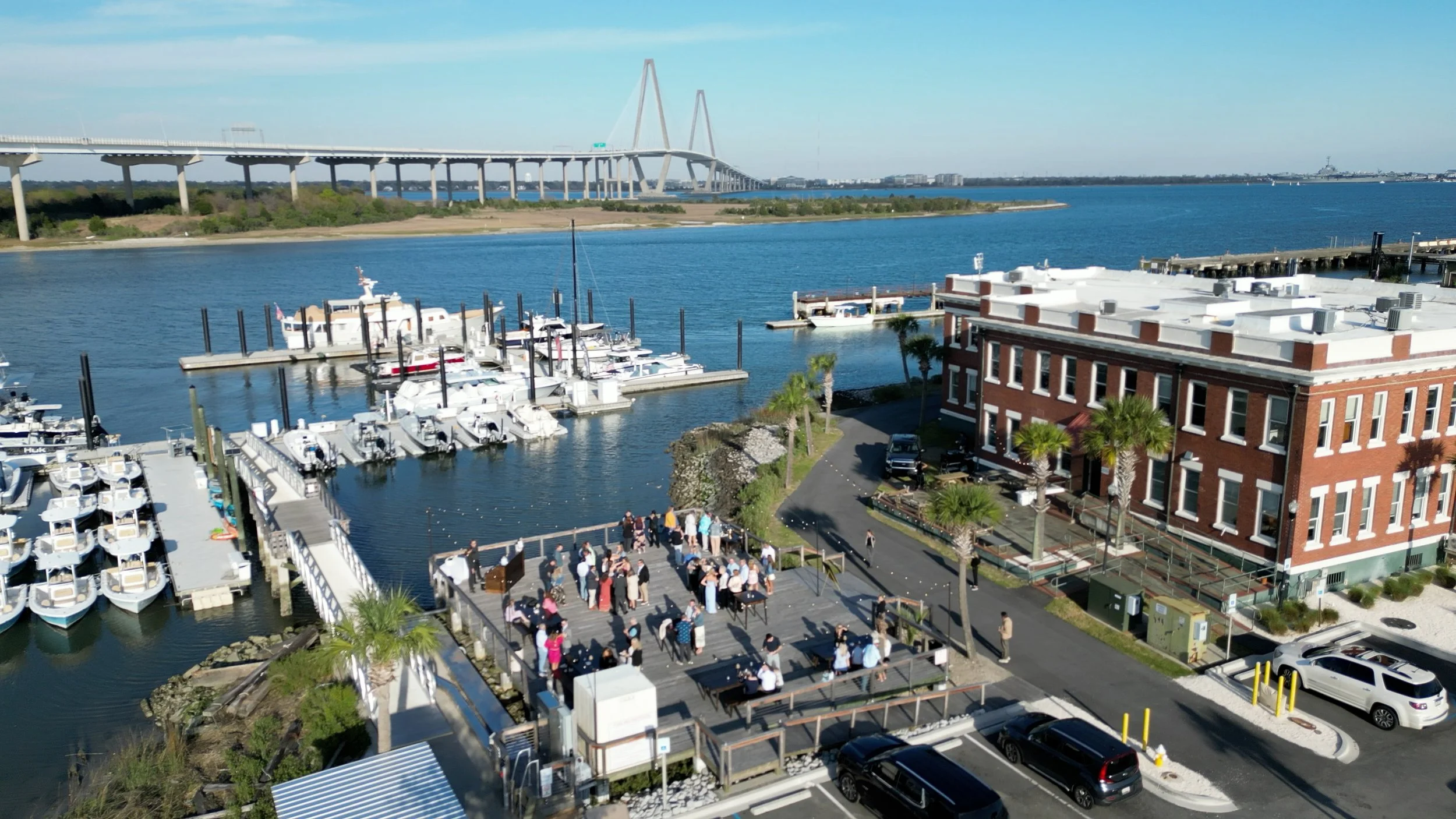 A marina with boats docked, a crowd of people gathered outside, and a large bridge in the background over a body of water.