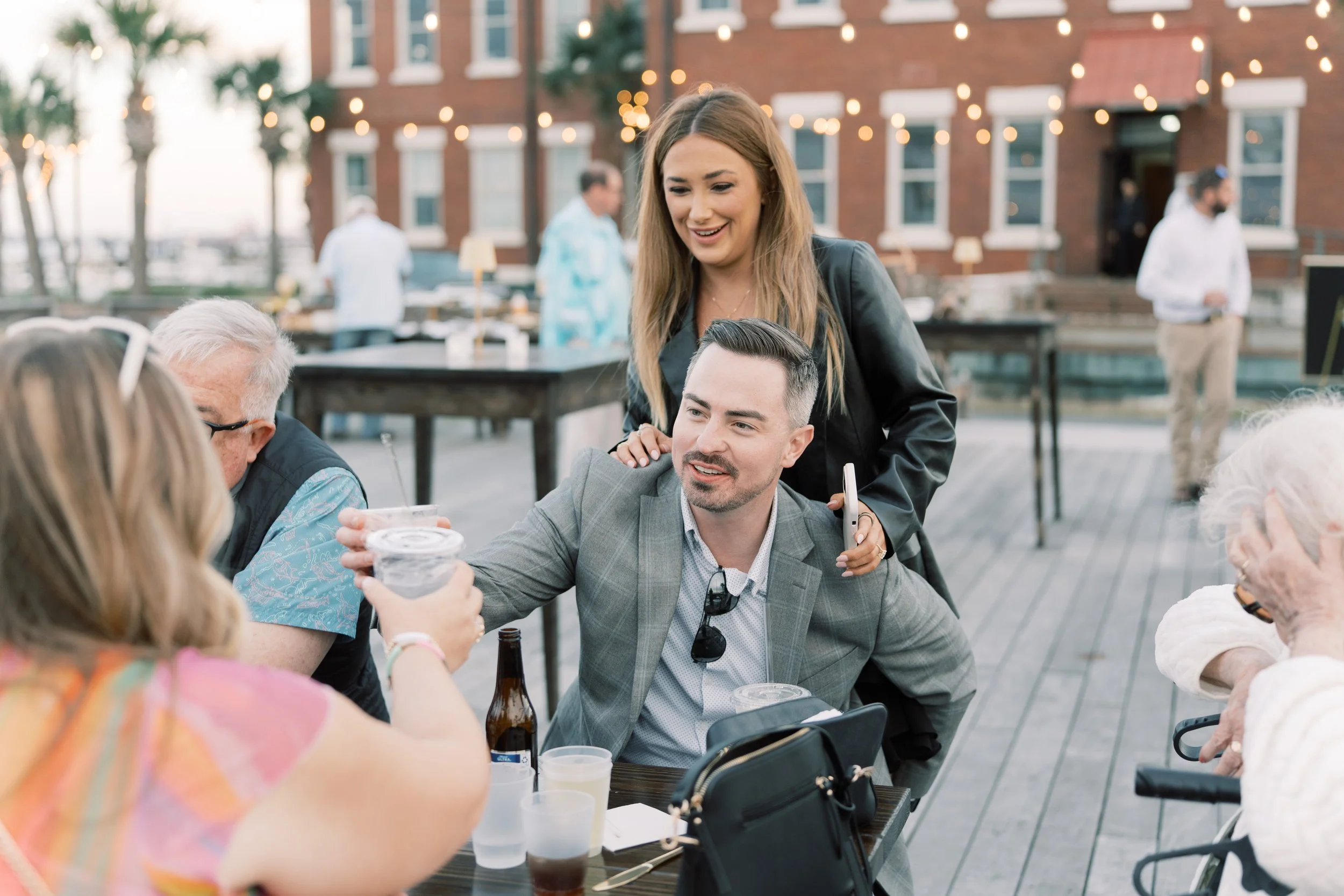 People enjoying drinks at an outdoor gathering on a patio with string lights and a brick building in the background.