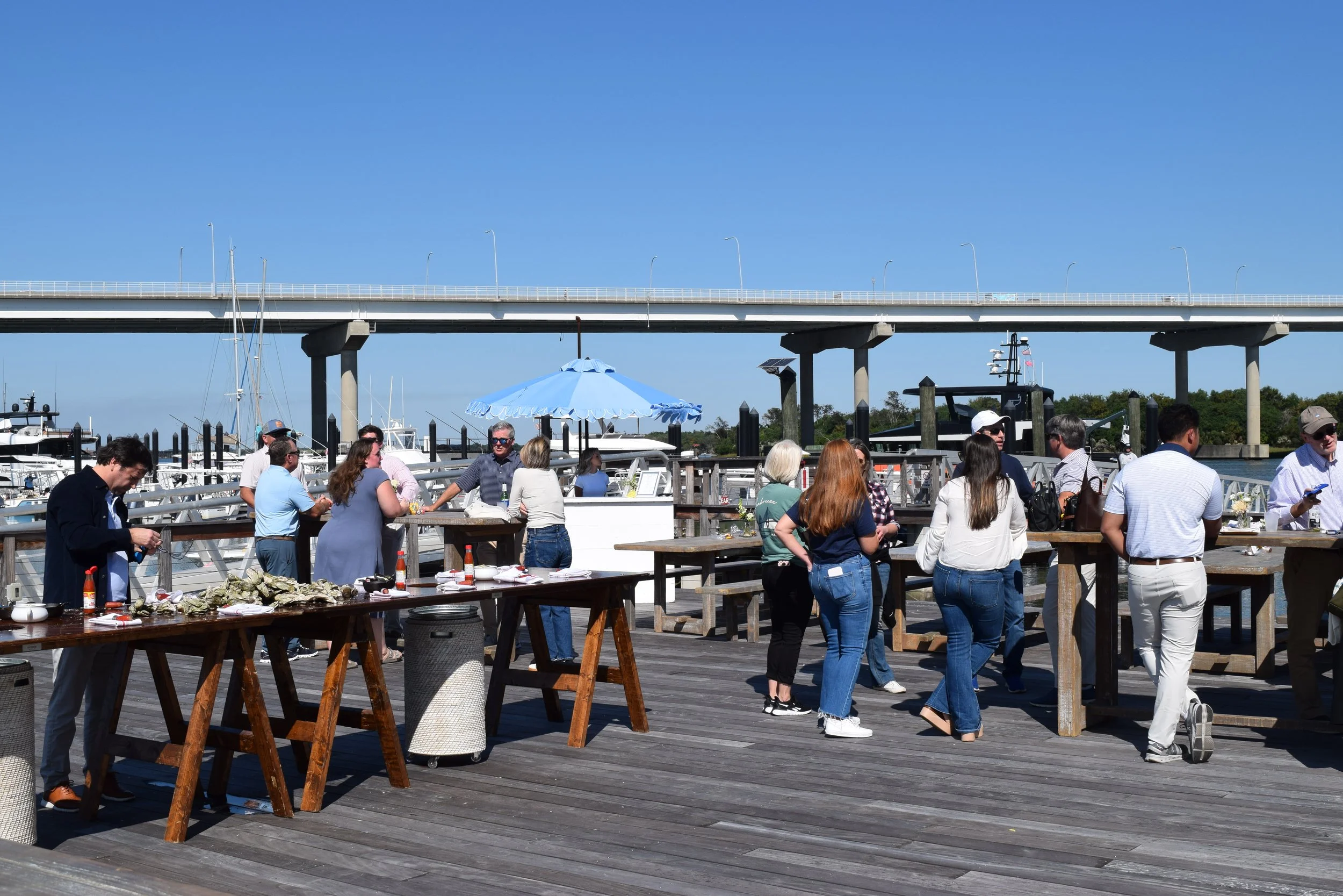 People gathered on a wooden dock for a seafood event, with boats docked nearby and a bridge crossing in the background under a clear blue sky.