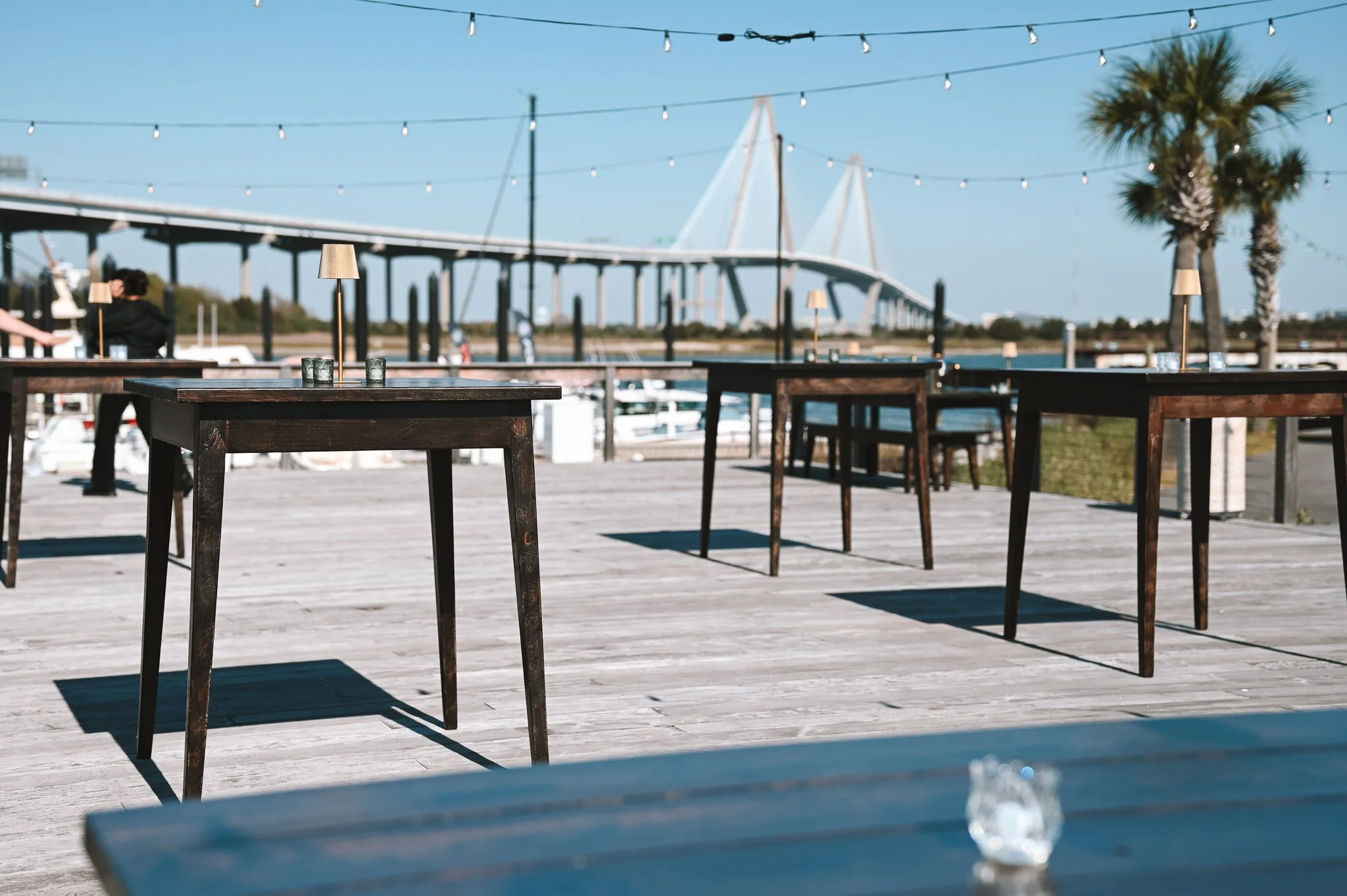 Outdoor patio with tables and lamps near a marina, with a bridge and palm trees in the background under a blue sky.
