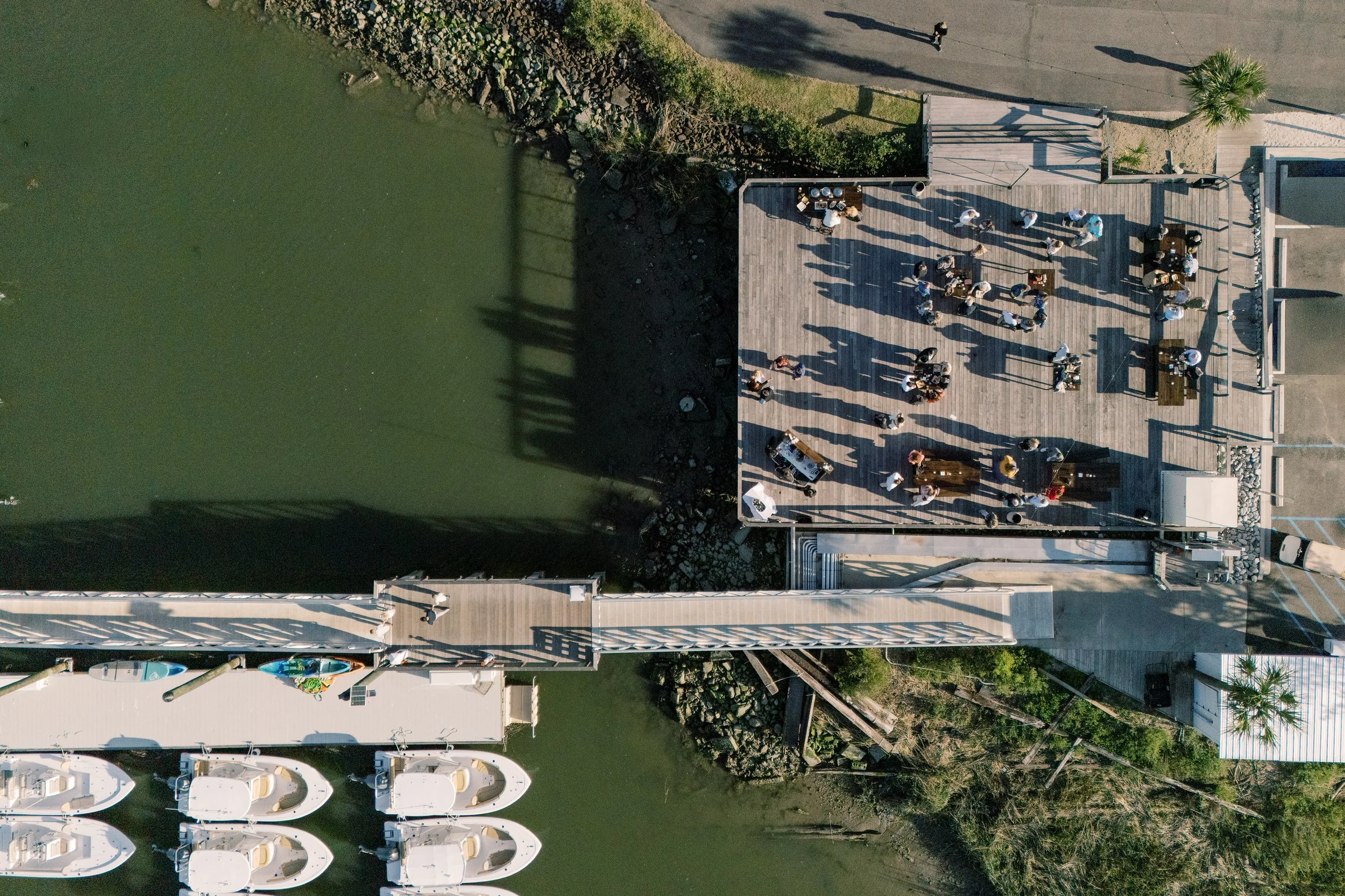 An aerial view of a waterfront restaurant or gathering area with people socializing on a wooden deck, boats docked along the water, and a adjacent road with pedestrians and a palm tree.