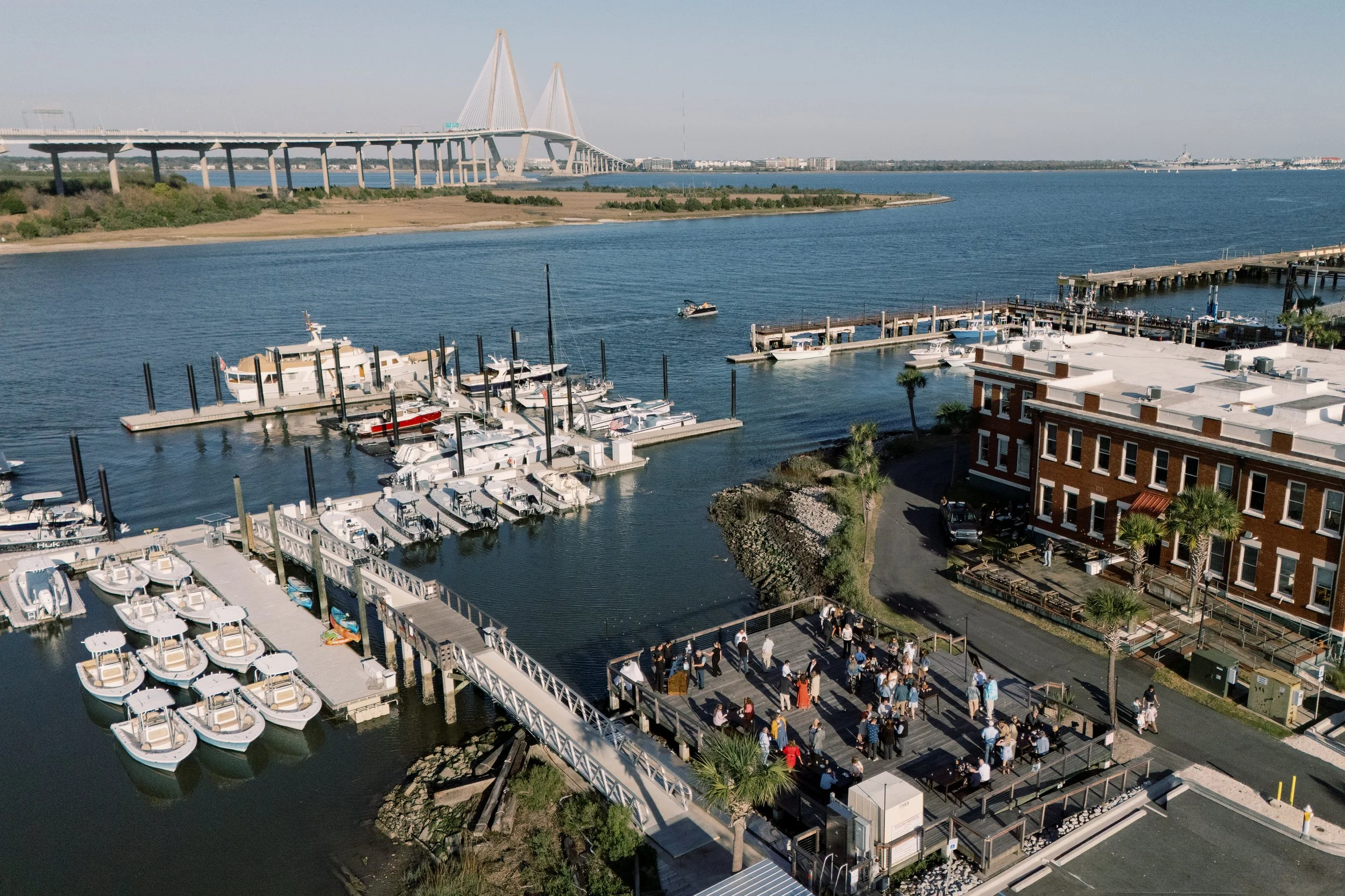 A marina with boats docked, a bridge in the background over a body of water, and a crowd walking along a path near the marina.