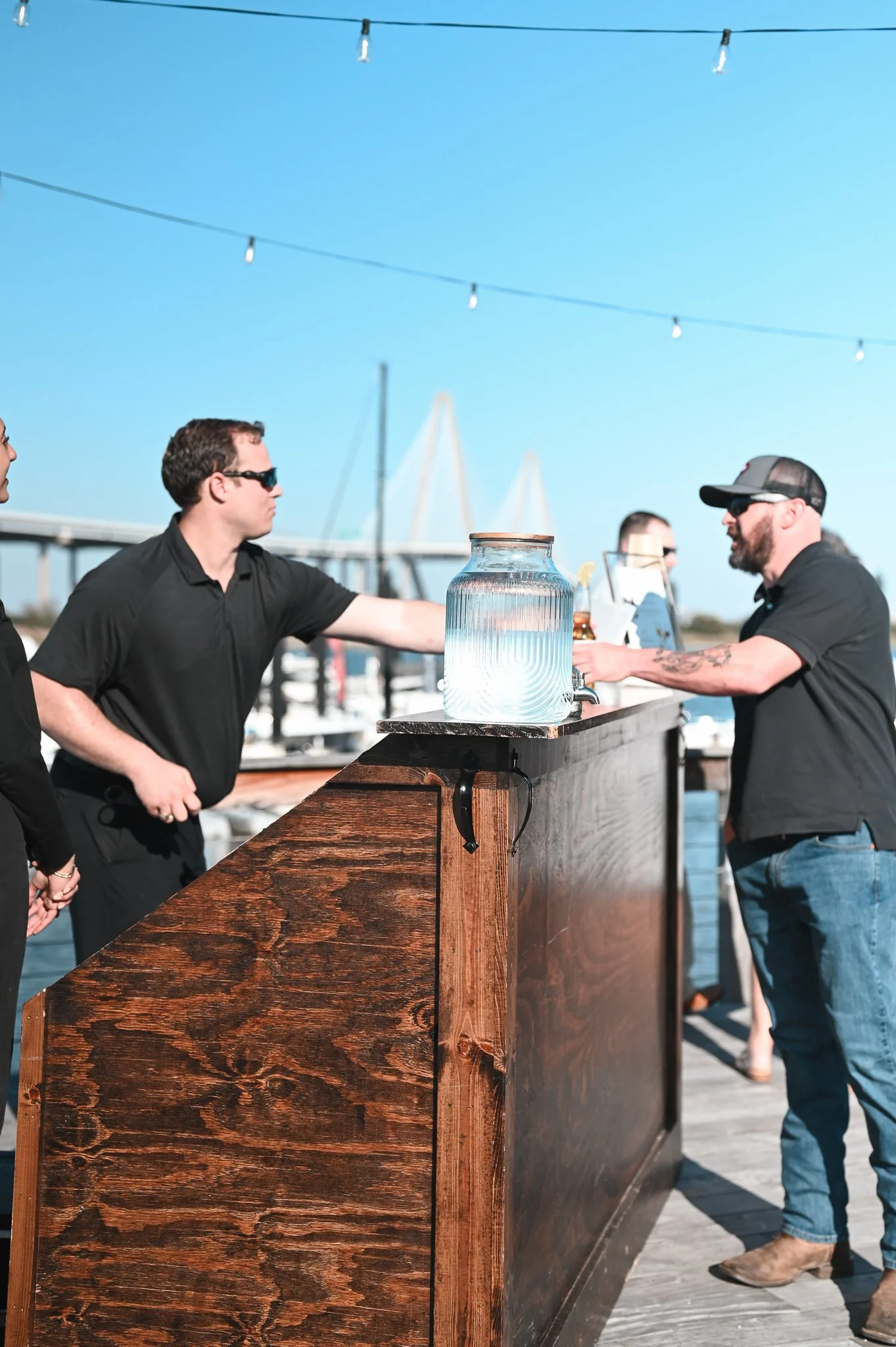 People socializing outdoors at a bar near the water, with a large jar of water on the bar, under string lights.