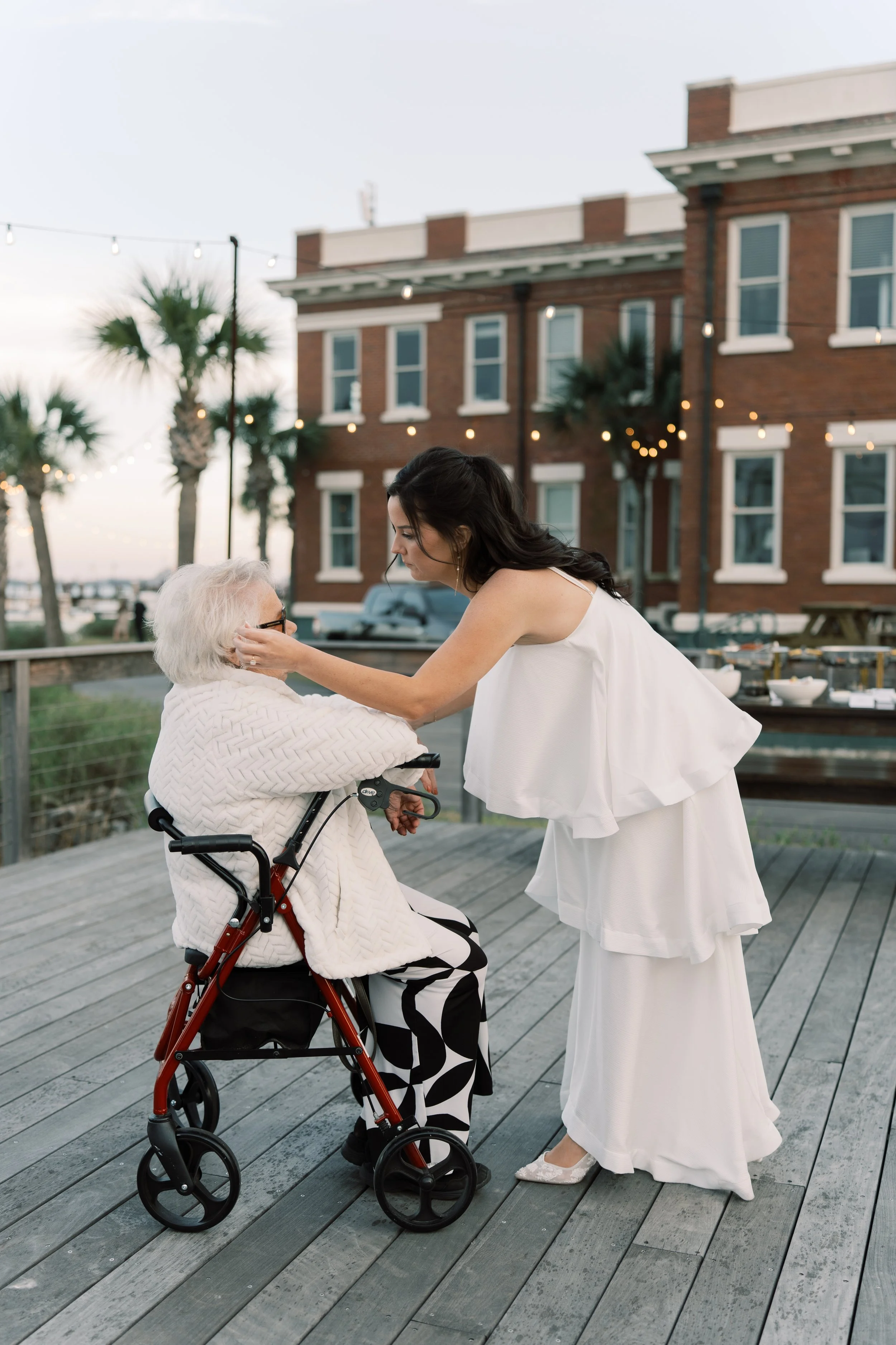 A young woman in a white dress is gently holding the face of an elderly woman in a wheelchair during sunset on an outdoor patio. There are string lights, trees, and red brick buildings in the background.