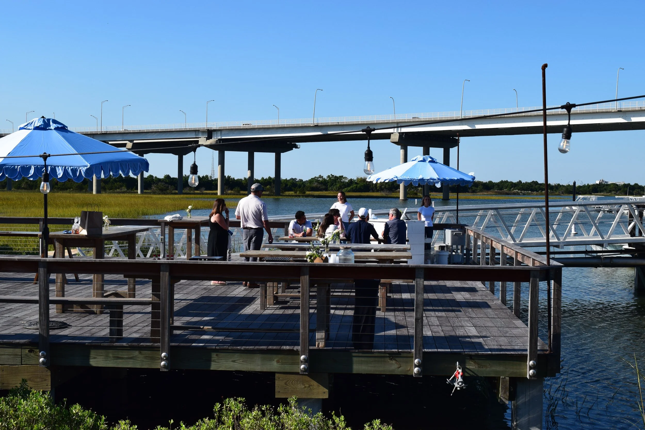People dining on a pier by the water with large blue umbrellas, a bridge in the background, and string lights hanging above.