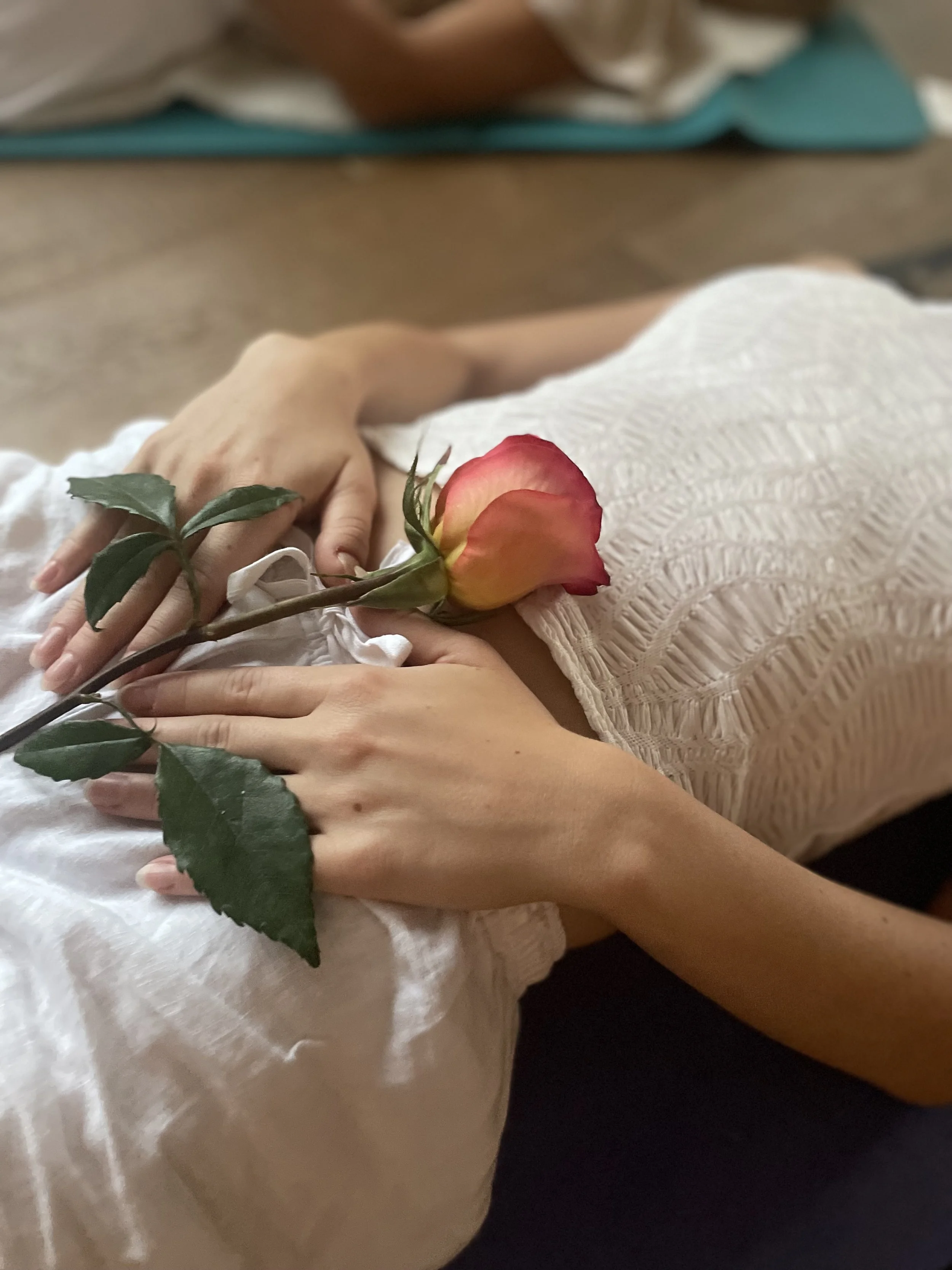 Eine Person liegt auf einem weissen Stoff, hält eine rosa und gelbe Rose auf ihrer Brust, während sie eine Hand auf die andere legt.