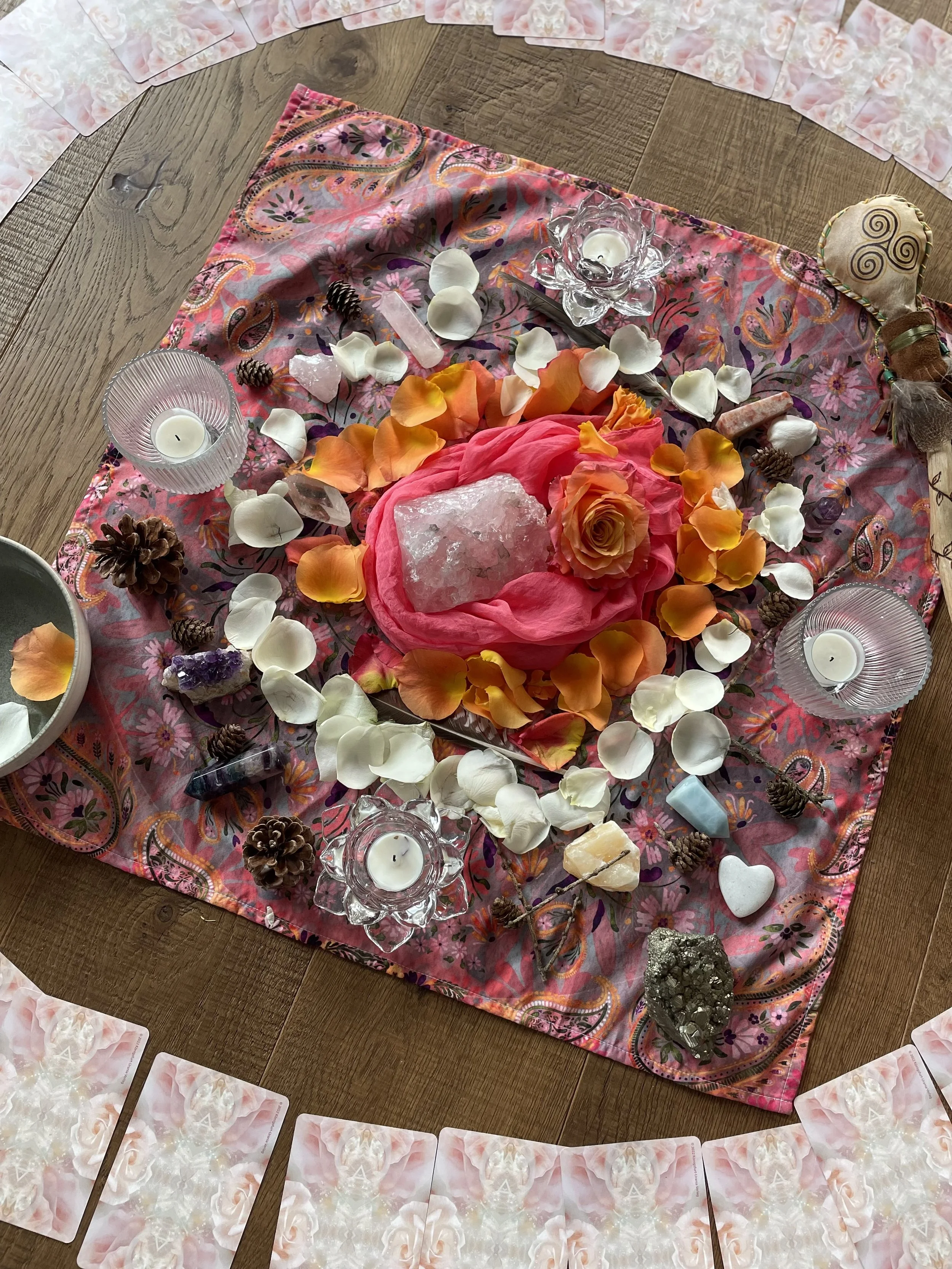 Eine Zeremonie auf einem pinkfarbenen Stoff mit Kerzen, Kristallen, Muscheln, Blättern, Piniensamen, Steinen und einer Rose in der Mitte, umgeben von Karten in einem halbkreisförmigen Muster.