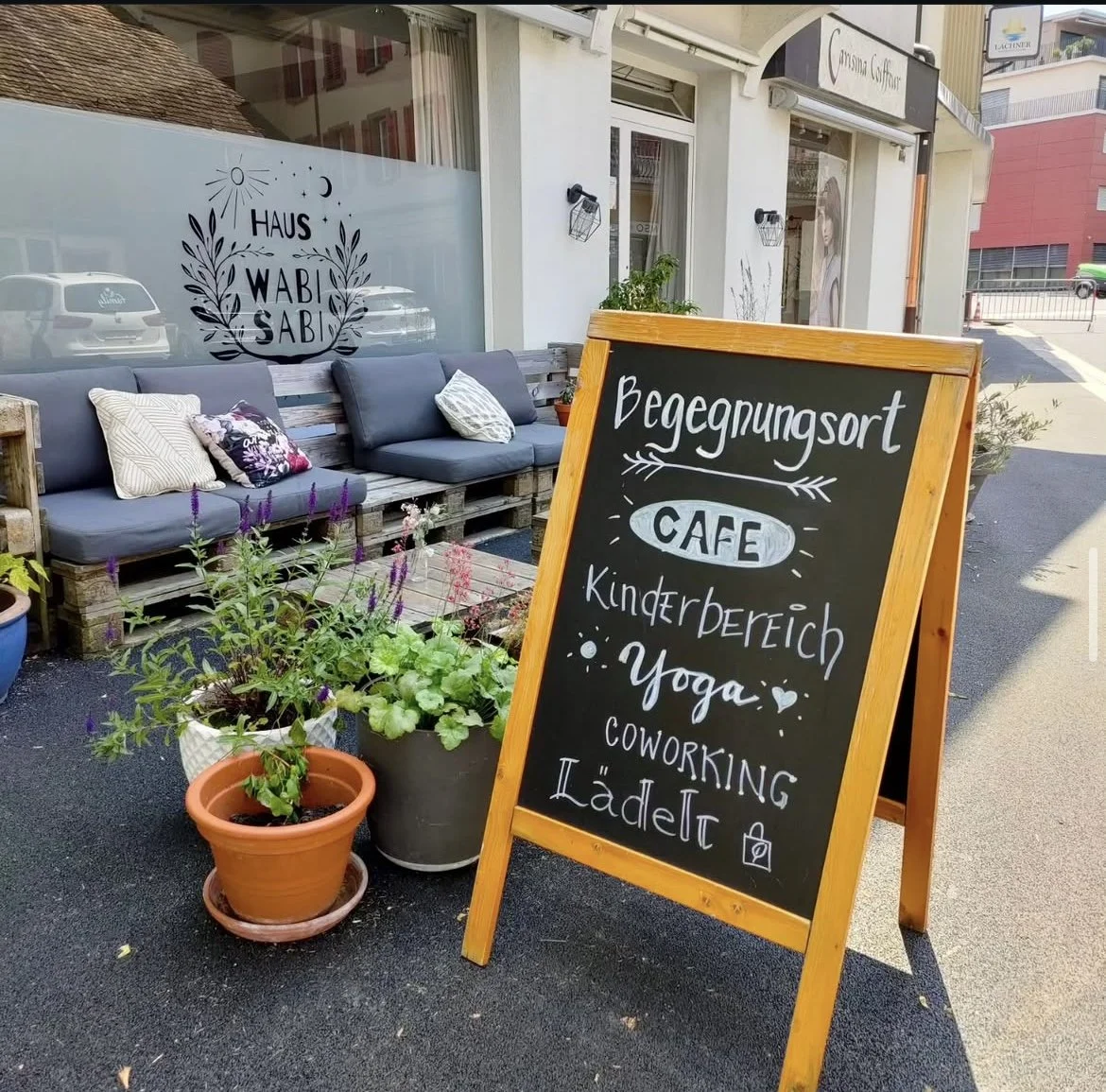 Strassenschild vor einem Café, Haus Wabi Sabi in Lachen, das Treffen, Yoga, Coworking und Lädeli anbietet, mit blumengesäumter Aussenfläche und Sitzbank.