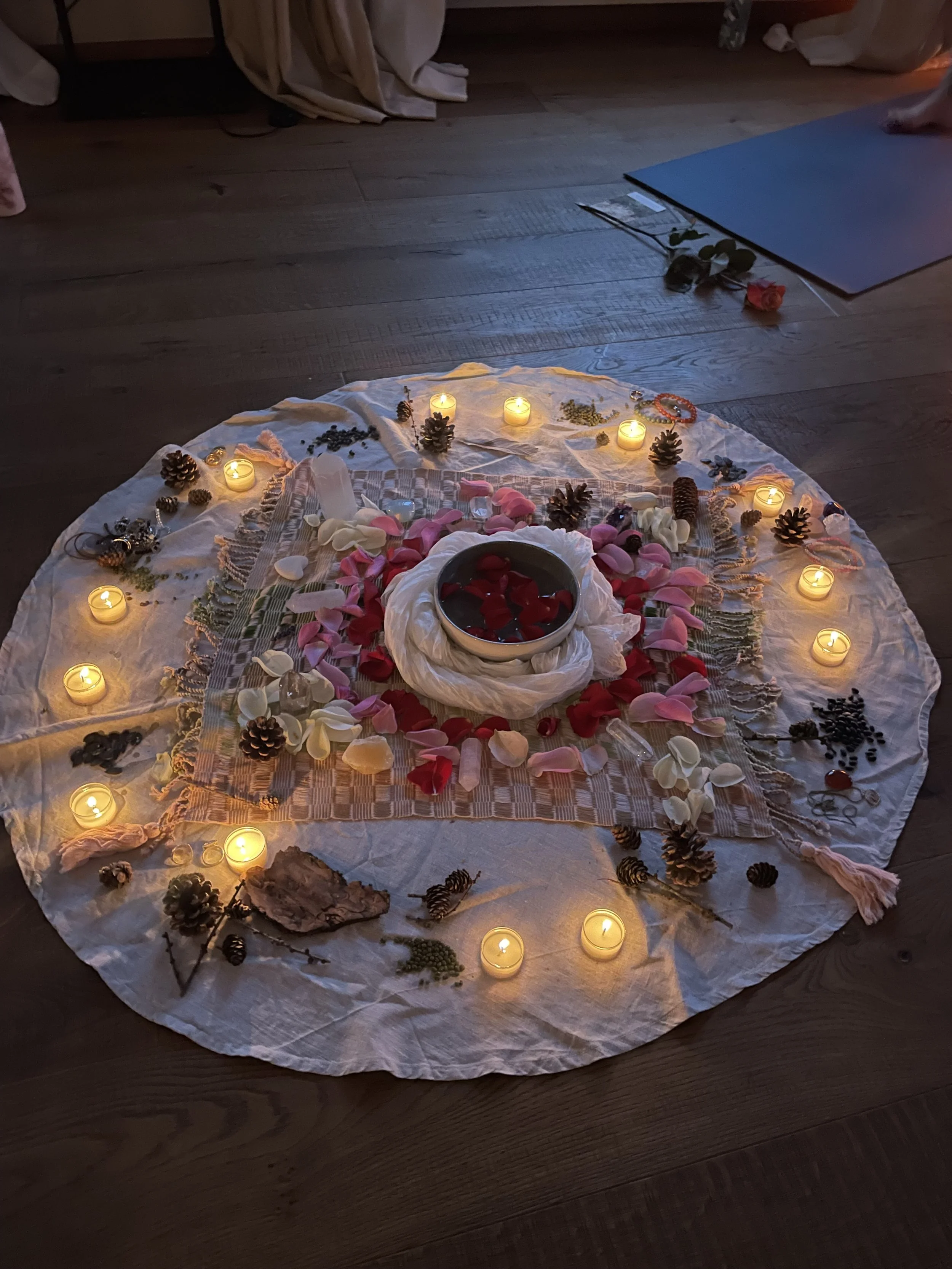 Eine Zeremonie aus Kerzen, Blumenblättern, Ästen und Steinen auf einem weissen Tuch, auf einem Holzboden.