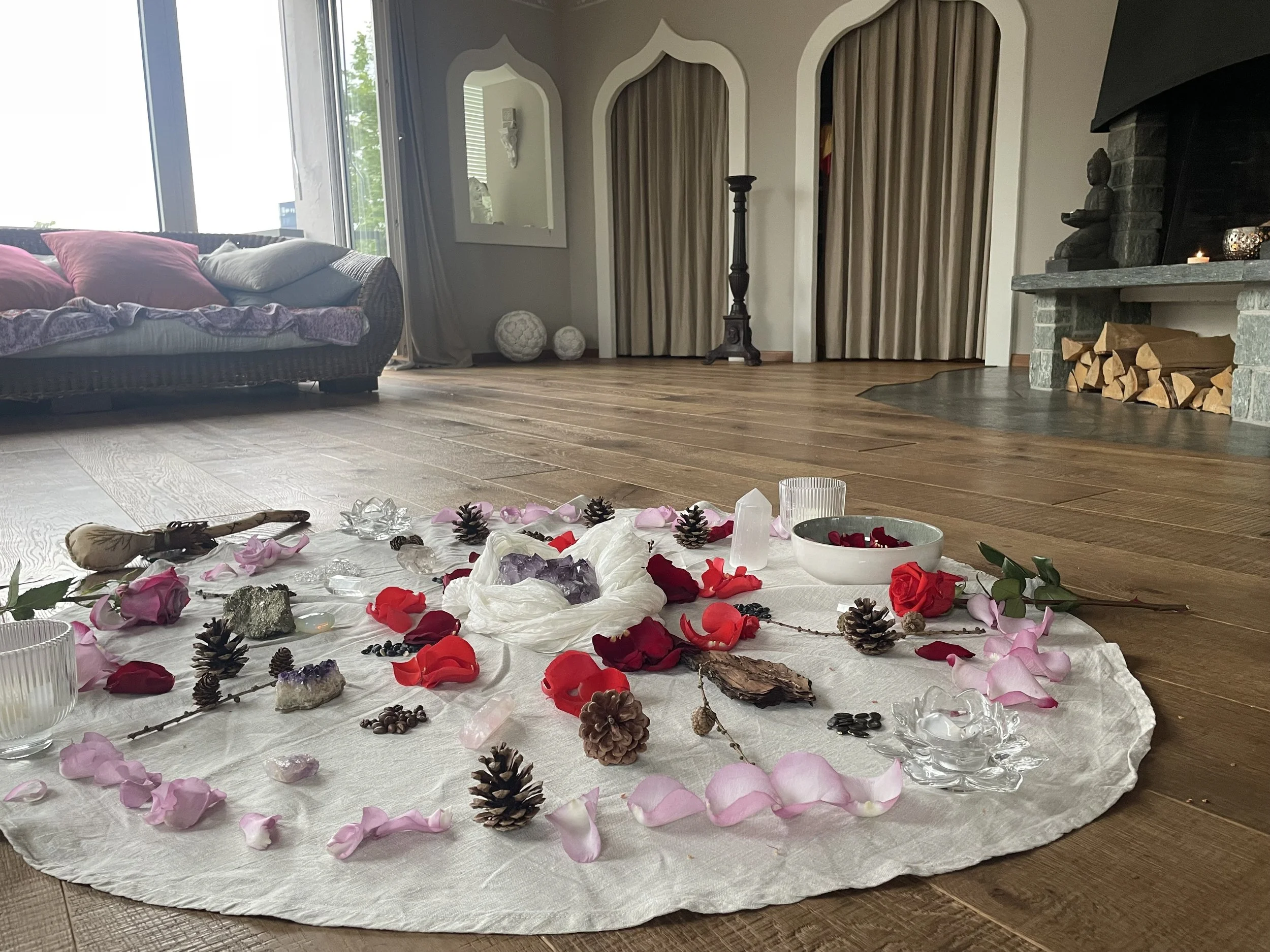Schossraum-Zeremonie mit Blumenblättern, Tannenzapfen, Kristallen und Steinen auf einem weißen Tuch auf einem Holzfussboden, Sessel, Kamin und Vorhängen.