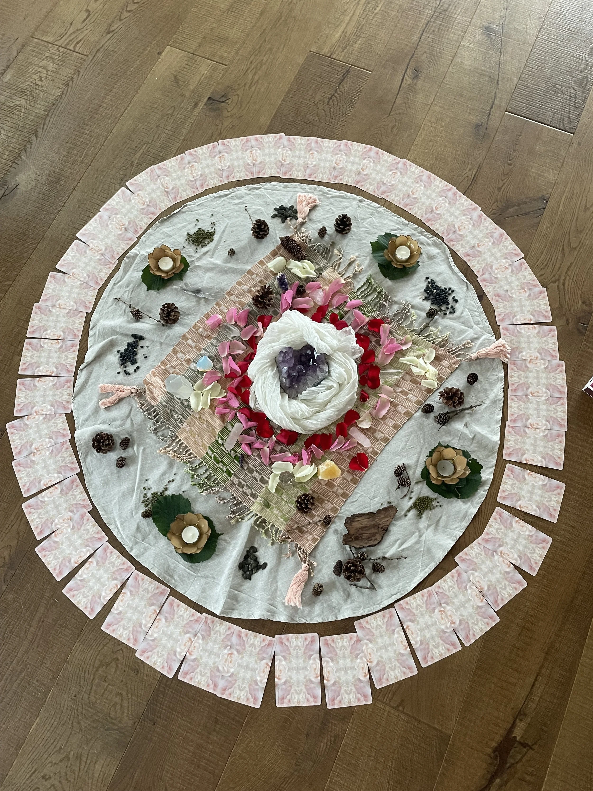 Bild mit Zeremonie aus Steinen, Blüten und Kerzen auf einem weissen Tuch, umrahmt von Karten auf einer Holzoberfläche.
