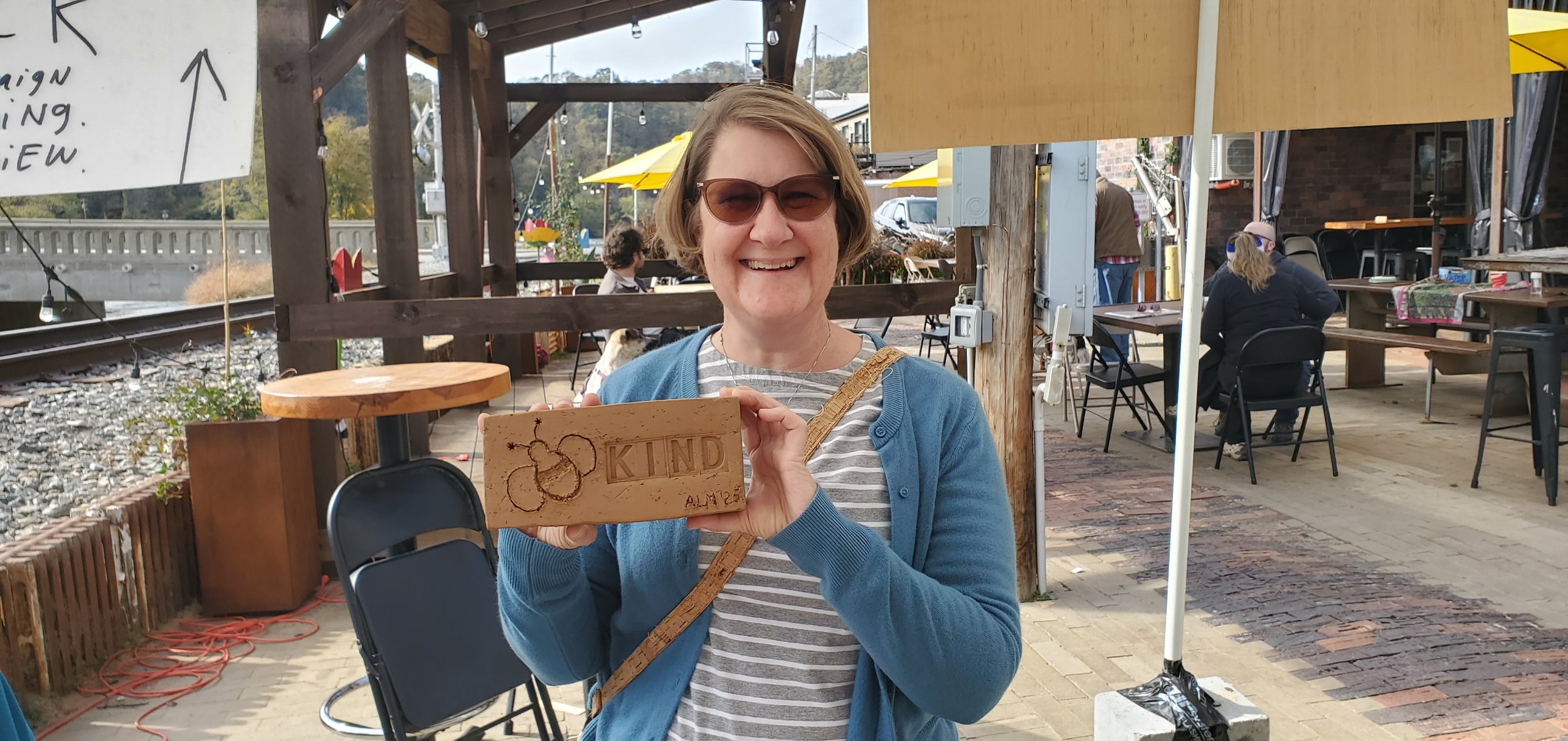 A woman with short light brown hair, wearing sunglasses, a striped shirt, and a blue cardigan, is smiling and holding a wooden block with the word 'KIND' carved into it and a drawing of a bee, outdoors at a cafe or restaurant with tables and chairs.