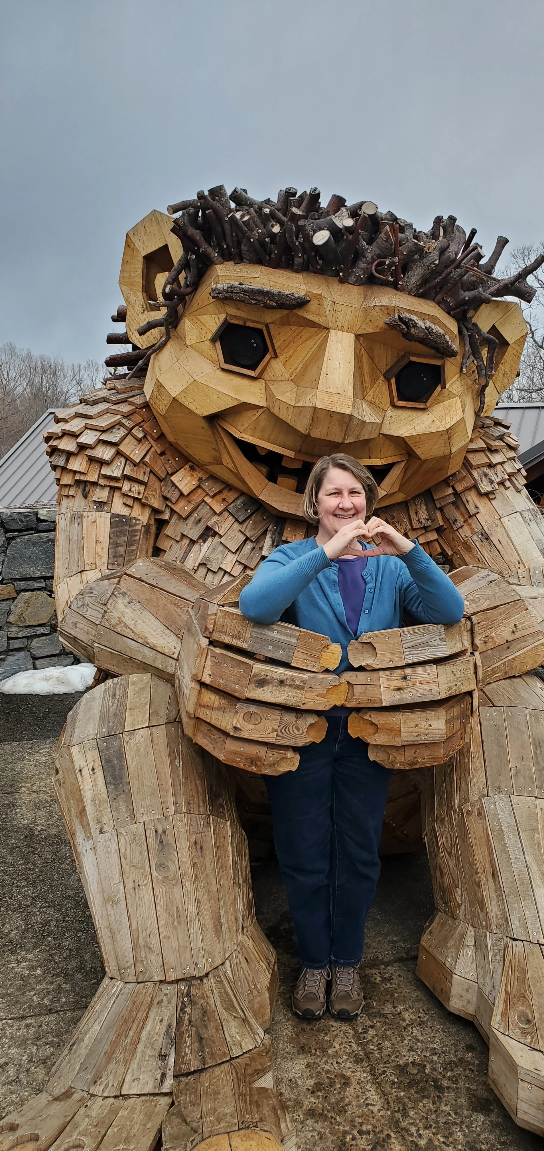 A woman standing inside a large wooden sculpture of a Dambo troll, making a heart shape with her hands in front of her chest.